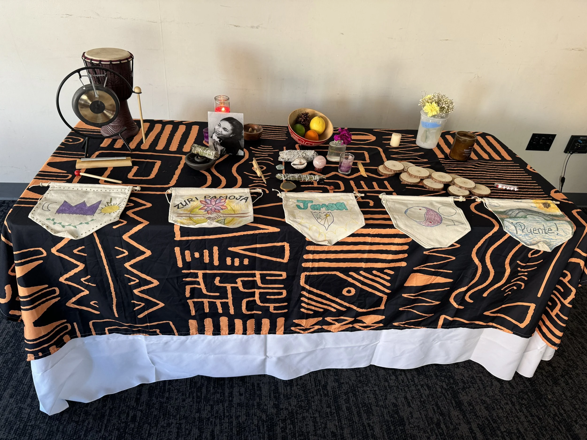A table covered with a black cloth with orange geometric patterns, displaying candles, a black and white photograph of a woman, handmade crafts, a fruit bowl, and decorative items. The table has a white cloth draped underneath.