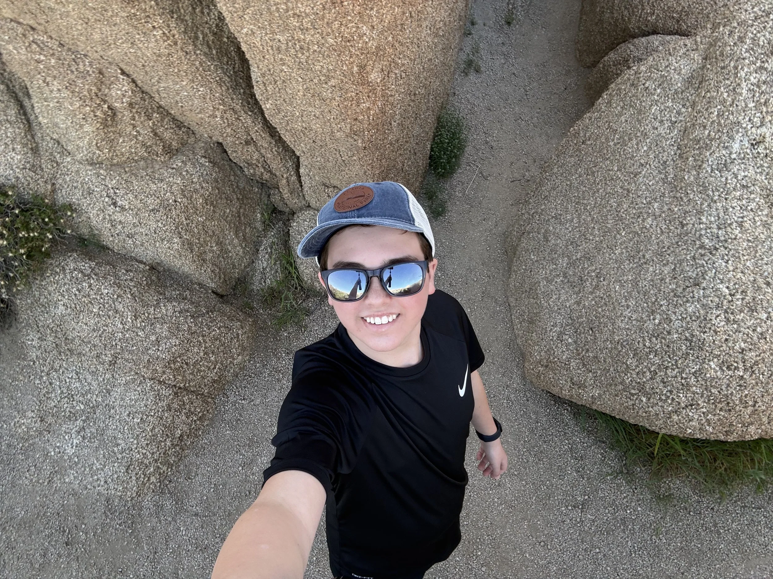 A person wearing sunglasses and a baseball cap taking a selfie in front of large rock formations on a sandy terrain.