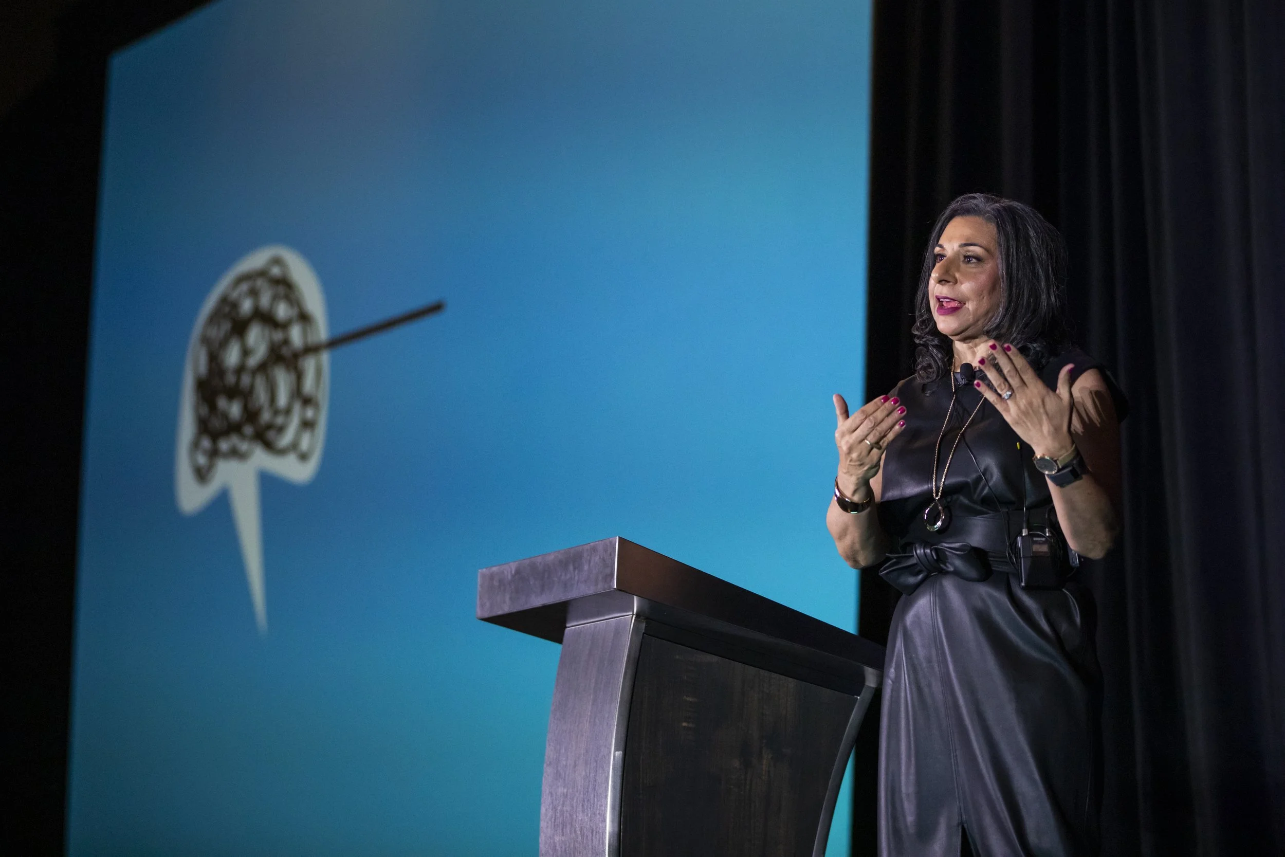 A woman with black hair and a black leather outfit speaking at a podium with a blue background showing a logo of a lion wearing a crown.