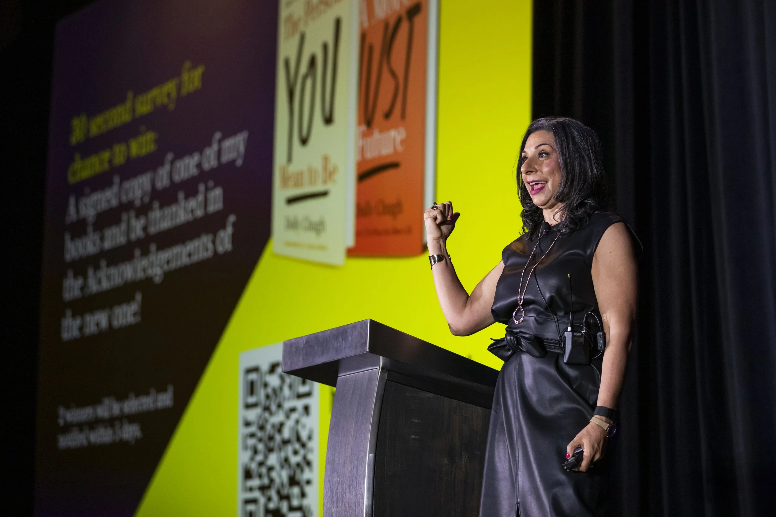 A woman with black hair and dark makeup, wearing a black leather dress with a bow on the side, speaking at a podium during a presentation or conference. Behind her is a large screen displaying colorful text and QR codes.
