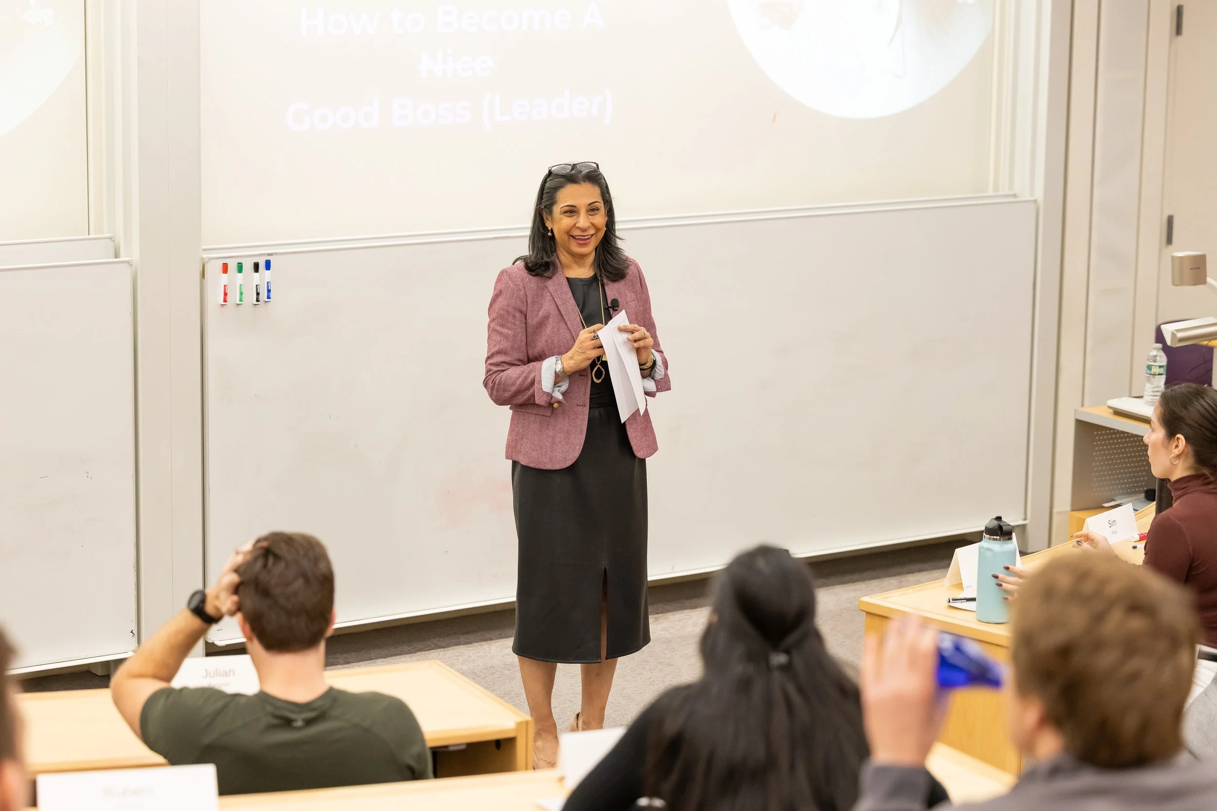Woman with dark hair and pink blazer presenting in front of a white board