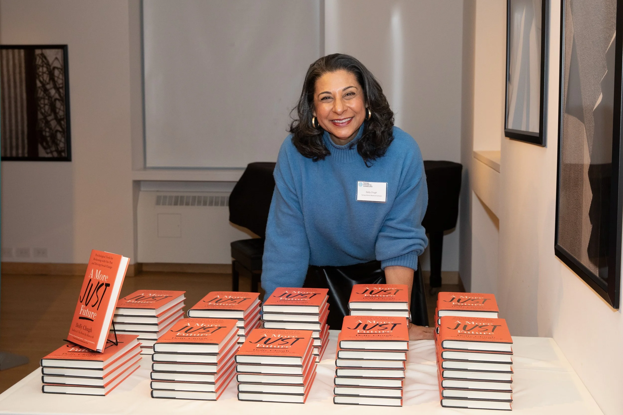 A woman with dark hair, wearing a blue sweater and gold hoop earrings, smiling while sitting behind a table covered with multiple stacks of orange and black books titled "A More Just Future" by Dolly Chugh at a book signing or event.