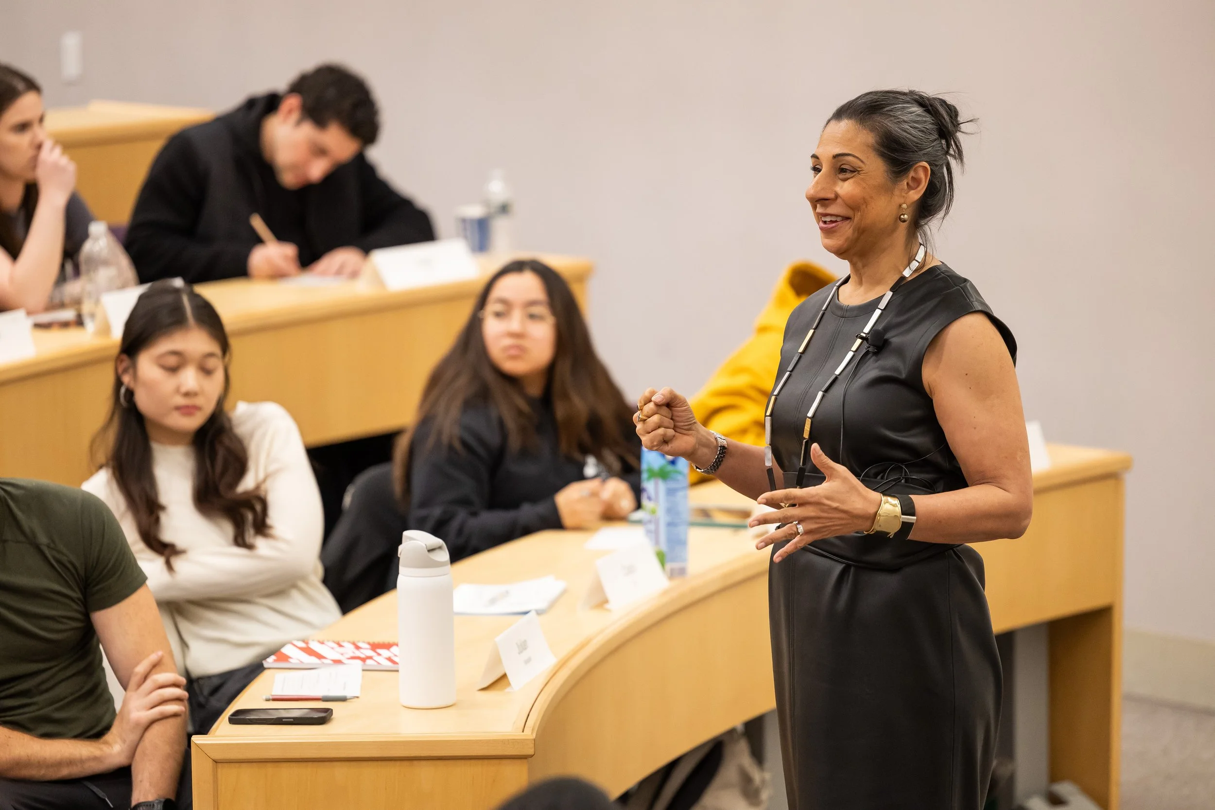 A woman with dark hair in a ponytail, wearing a black sleeveless top and a necklace, stands and speaks to a group of people seated at table in a classroom or conference room.
