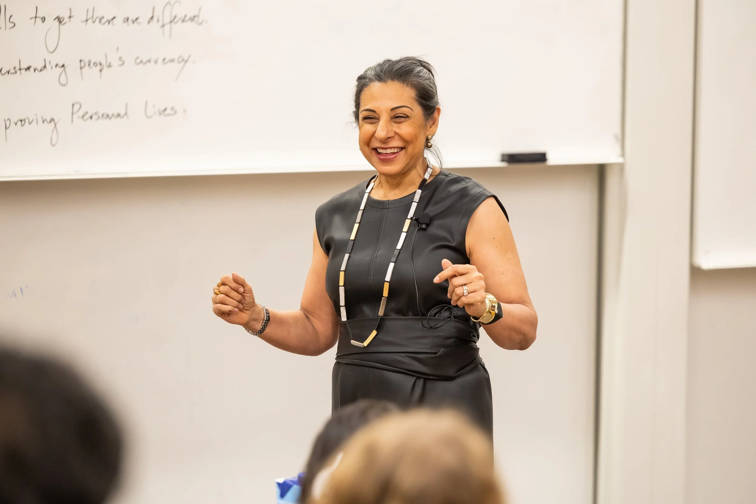 A woman with dark hair tied back, wearing a sleeveless black dress and a large beaded necklace, is smiling and speaking in front of a whiteboard in a classroom setting.