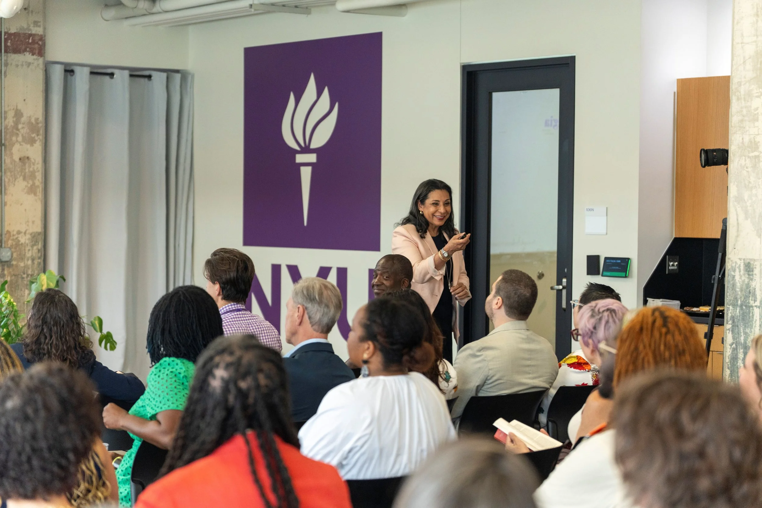 A diverse group of people attending a conference in a modern room. A woman in a light-colored blazer is standing and speaking to the audience. There are large purple banners with white torch symbols and the word 'NYL' in the background, along with a door, a black camera, and some plants.