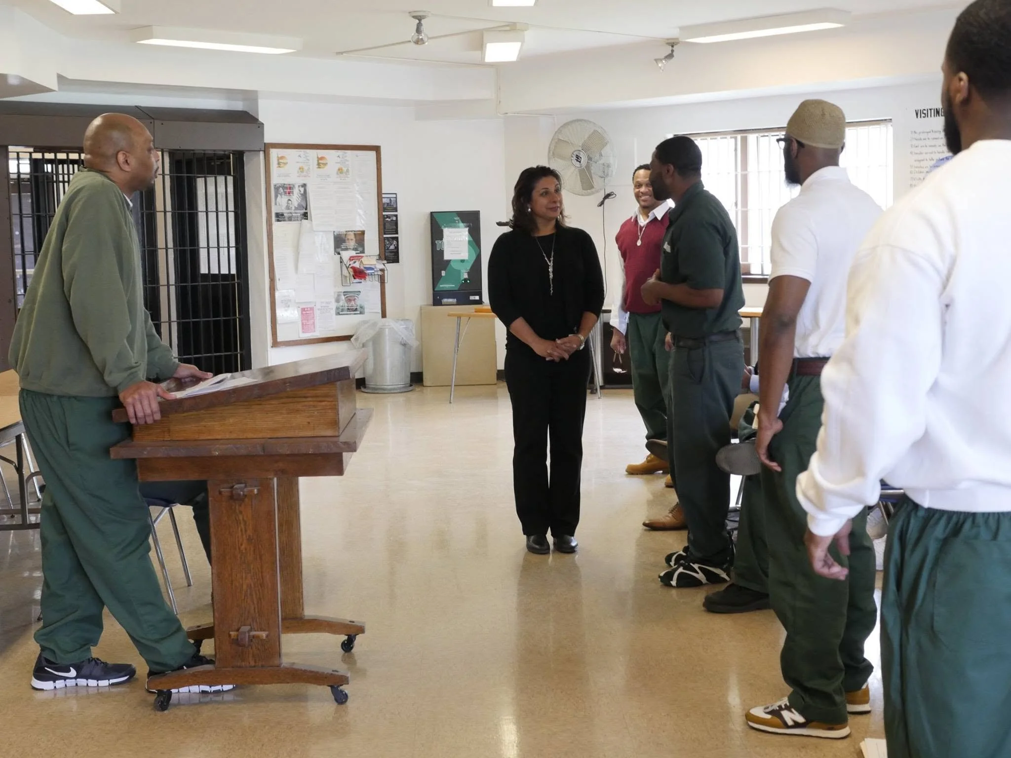 A group of six people standing in a line inside a room with white walls, engaged in conversation. The room has windows, a bulletin board with notices, and a fan mounted on the wall. One person, standing behind a wooden podium, is listening to the others, who are speaking or waiting their turn.