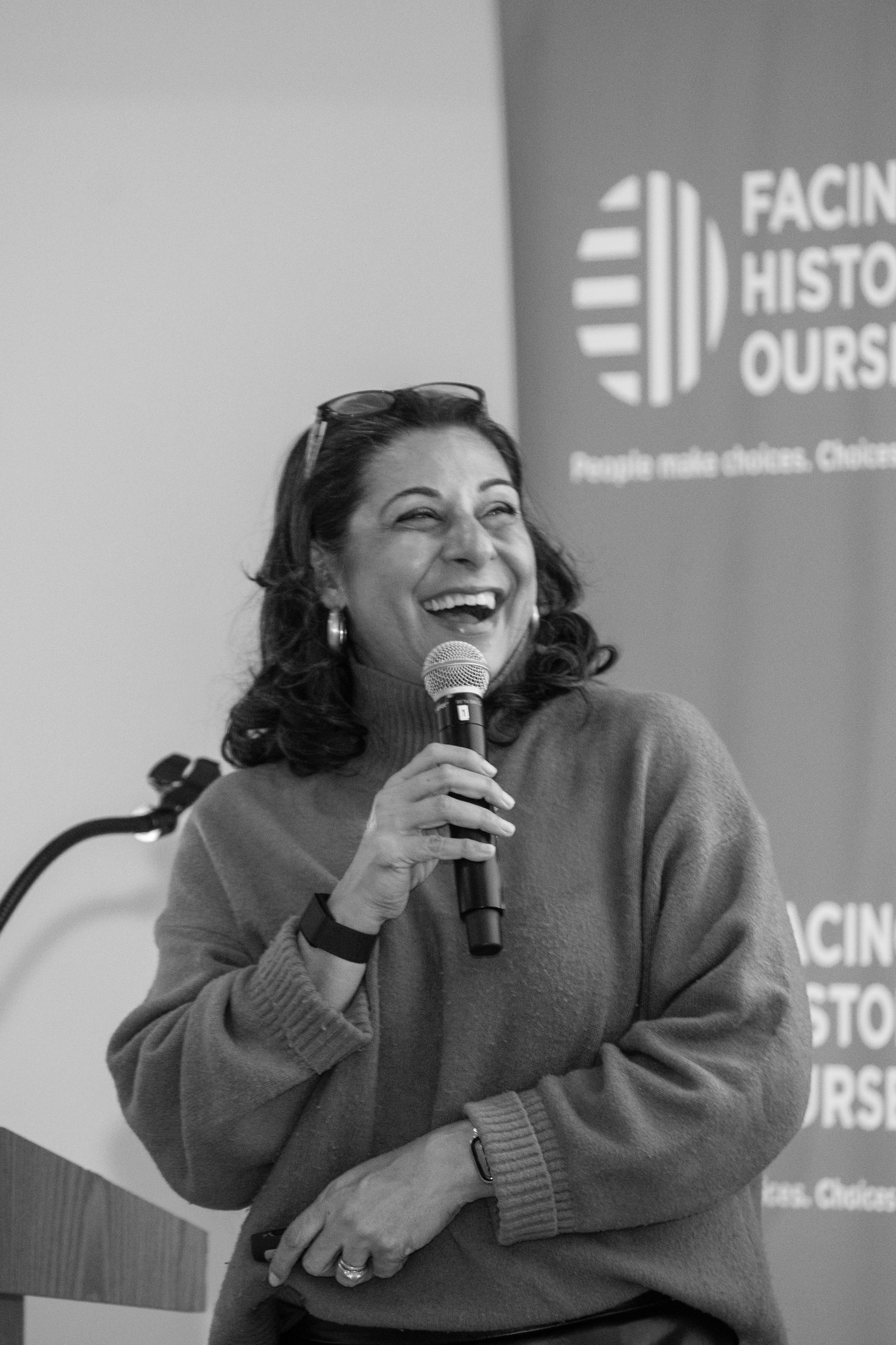 A woman smiling and speaking into a microphone at an event, with a banner in the background that reads 'Facing History Ourselves'.