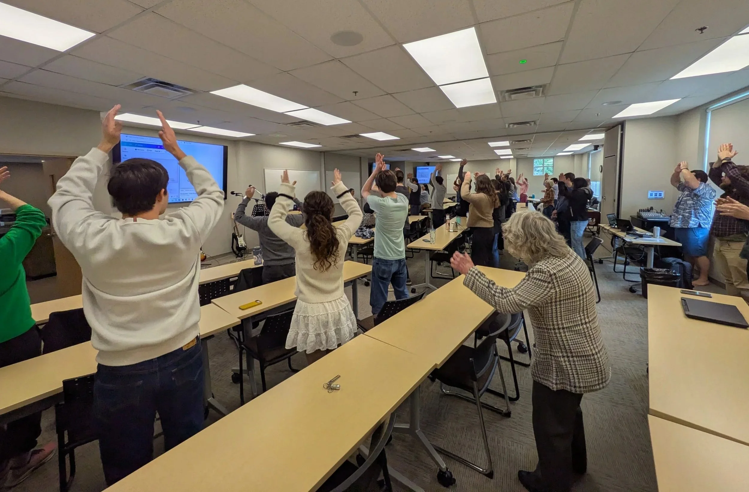 Group of people standing in a classroom with their hands raised, facing the front where a large screen displays blue content. Some individuals are taking notes, and there are tables and chairs arranged in rows.