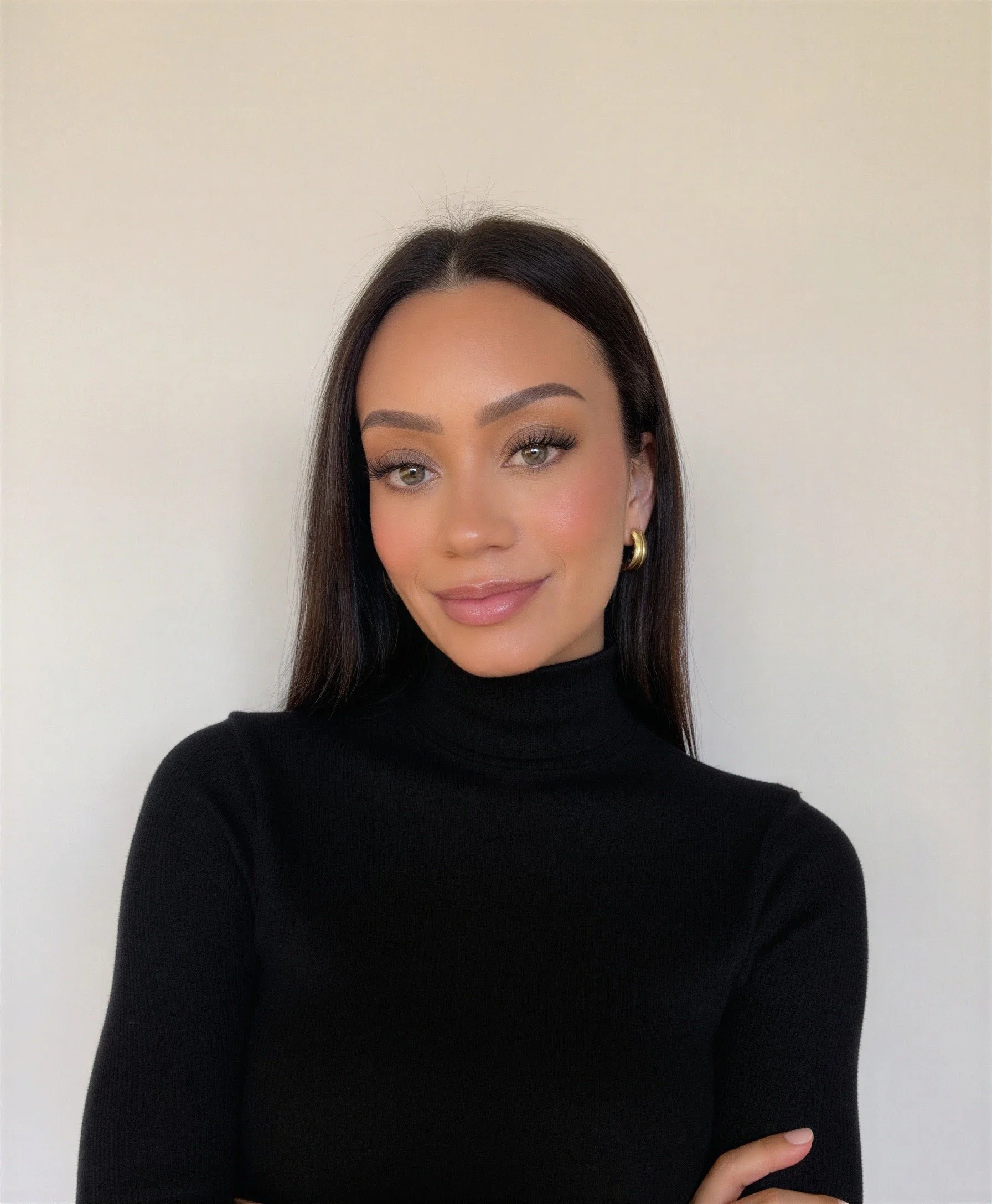 A woman with long dark hair and green eyes smiling, wearing a black turtleneck sweater and gold earrings, standing against a plain white wall.