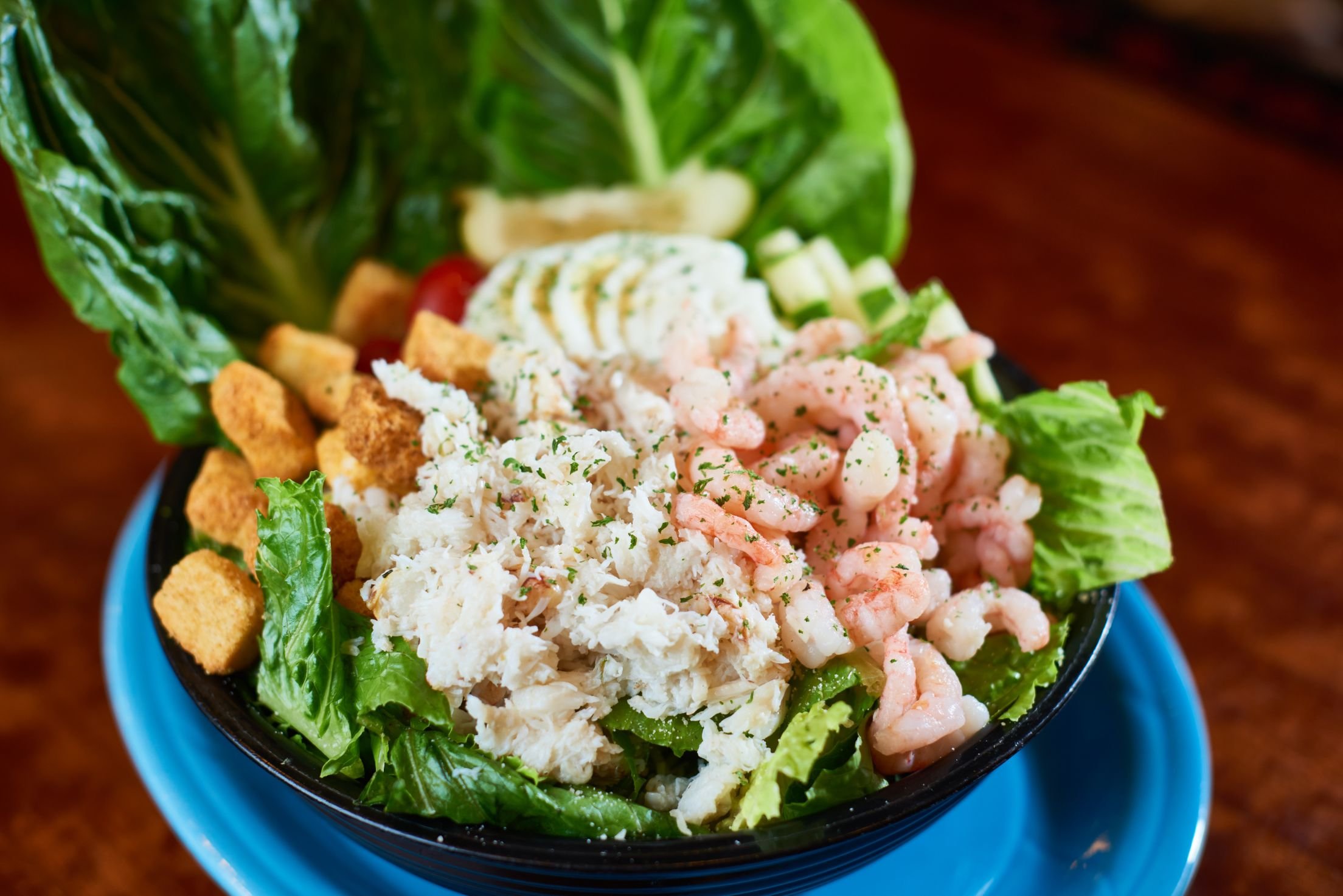A bowl of chopped leafy lettuce topped with shredded crab, diced cucumbers, chopped green onions, sliced cherry tomato, a scoop of potato salad, and croutons, on a wooden table.
