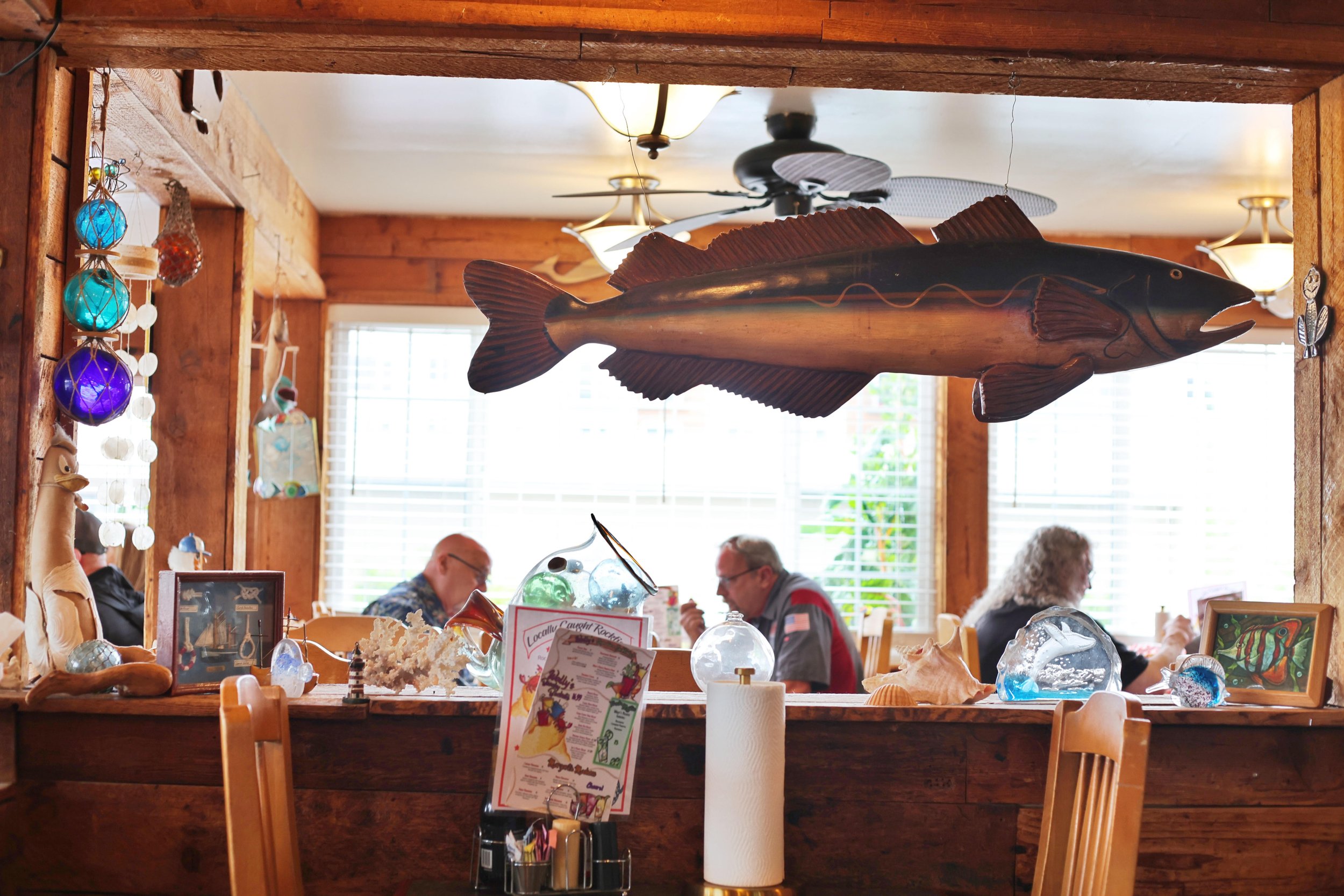 Inside a restaurant with wooden decor, a large fish sculpture hangs from the ceiling, and there are colorful glass buoys hanging on the side. Several people are dining, and sunlight streams through large windows.