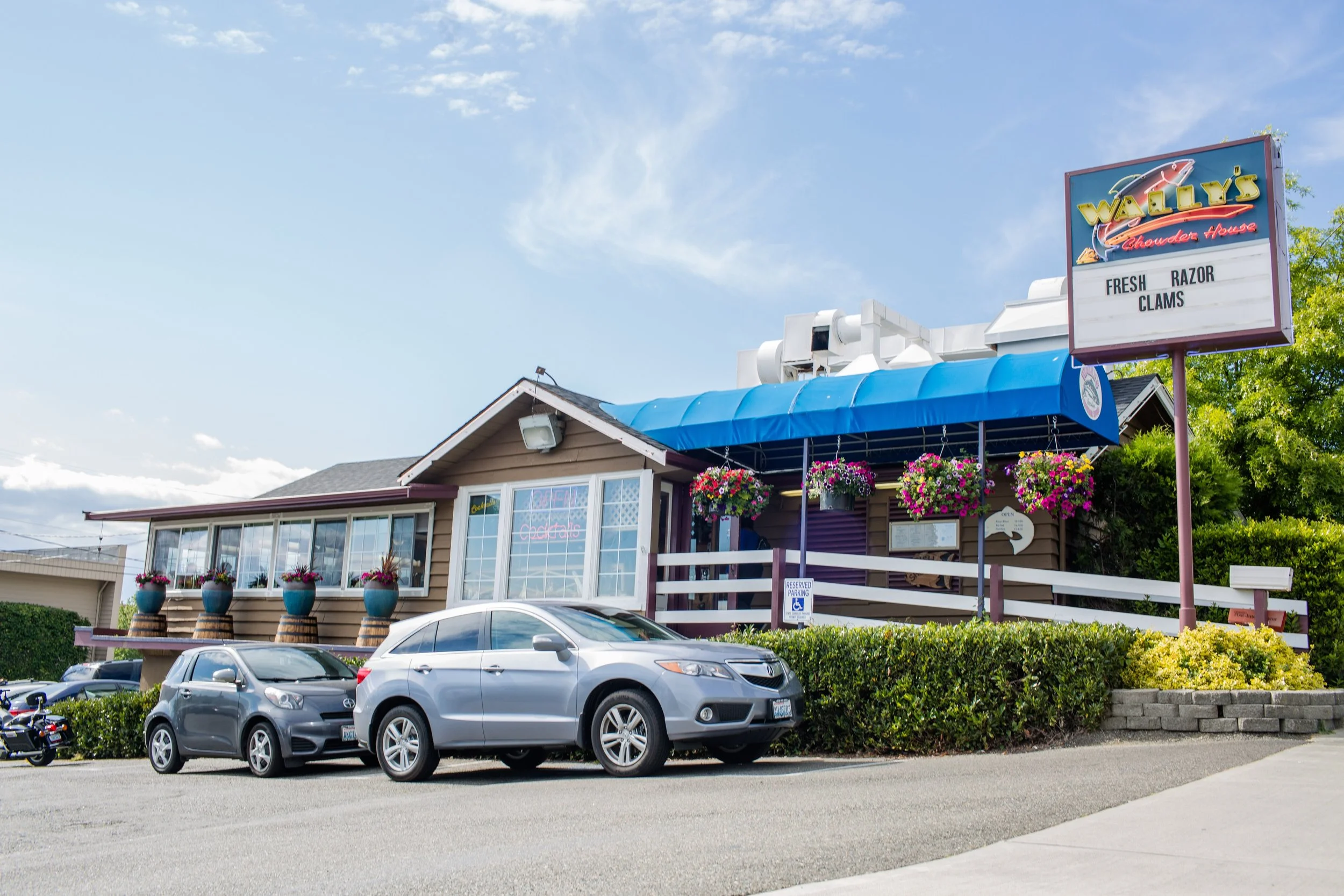 Exterior view of Wally's Chowder House restaurant with a parking lot in front, cars parked, and a sign displaying 'Fresh Razor Clams' against a partly cloudy sky.