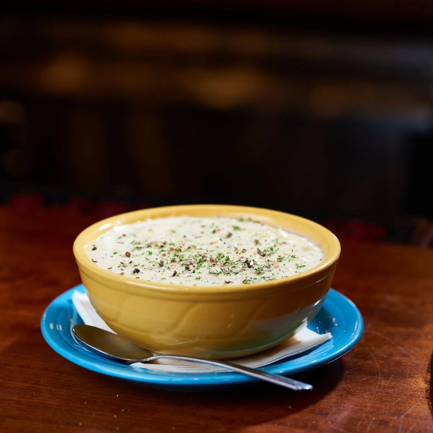 A bowl of creamy clam chowder garnished with herbs and black pepper, placed on a blue plate with a spoon, on a wooden table.