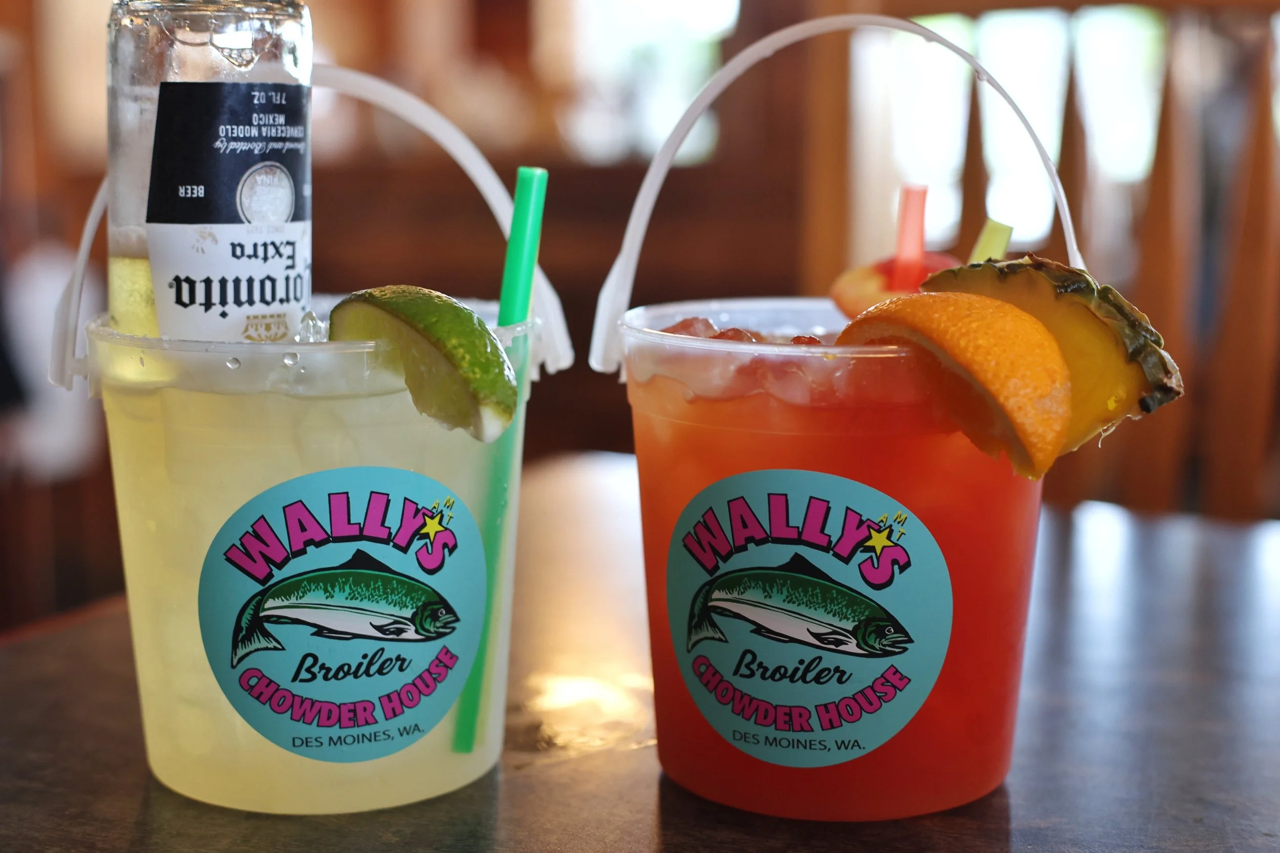 Two colorful drinks in large plastic buckets with straws, on a table, both topped with fruit slices and branded with Wally's Chowder House logo.