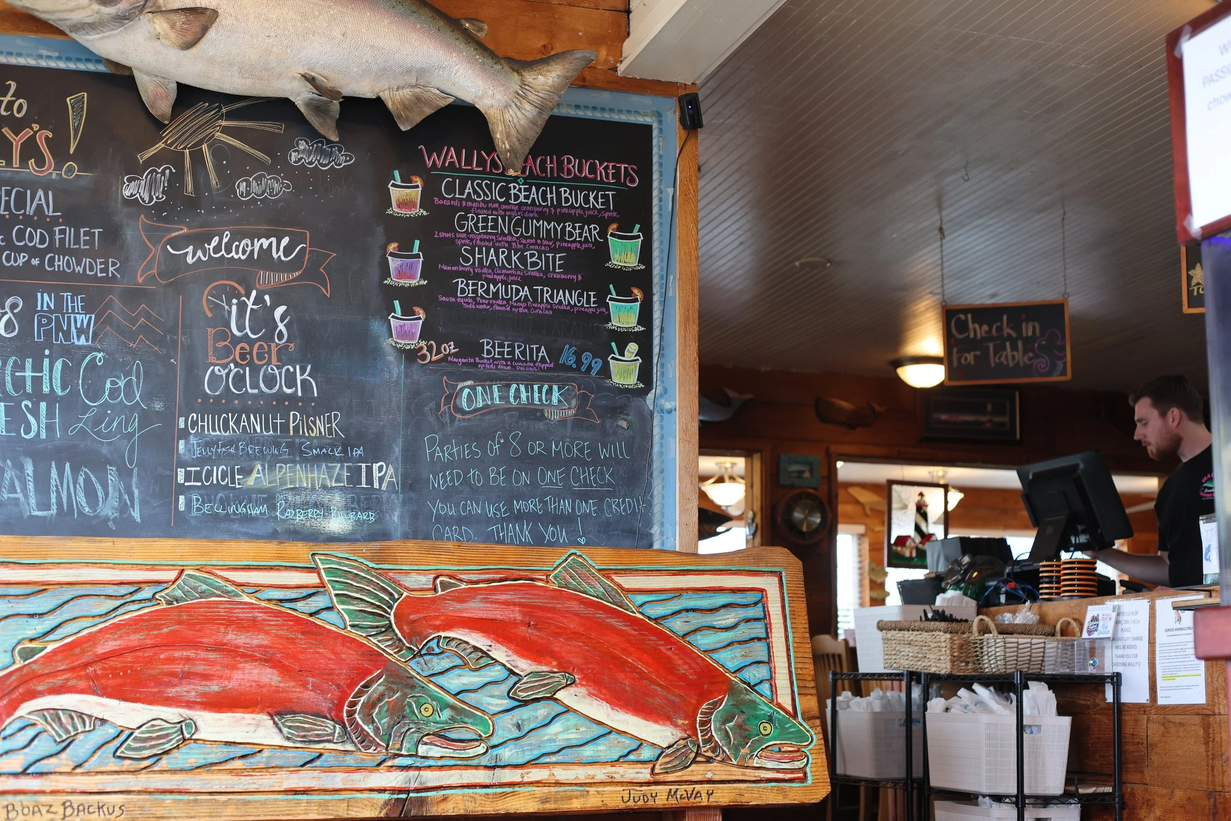 A chalkboard menu in a restaurant or bar with colorful writing listing various drink options, including classic beach bucket, green gummy bear, sharkbite, Bermuda triangle, and berita, with descriptions and prices. There is a mounted fish above the c