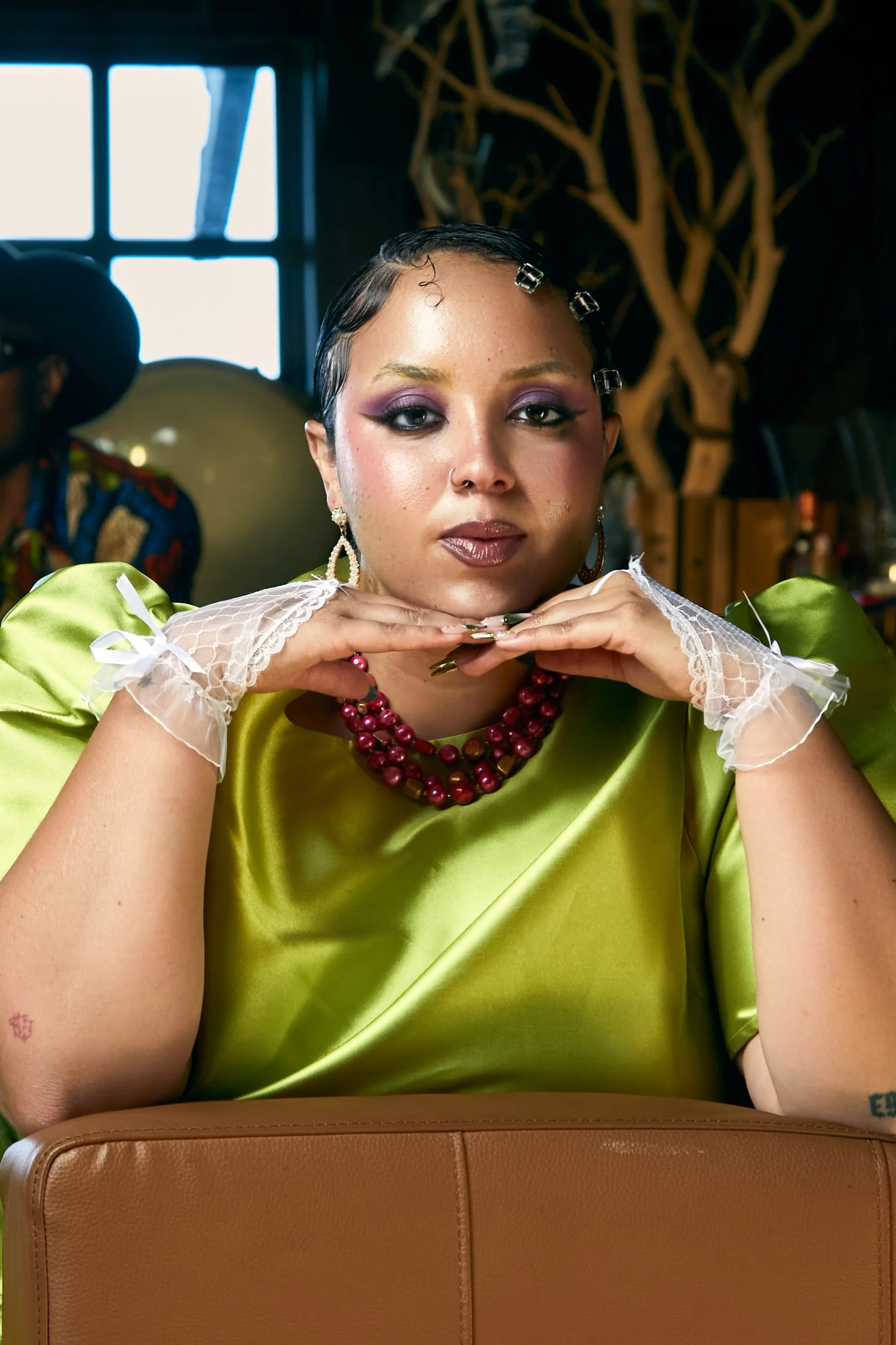 A woman with short dark hair, wearing purple makeup, pearls, and a shiny green dress, resting her chin on her hands in a room with a dark background and window light.