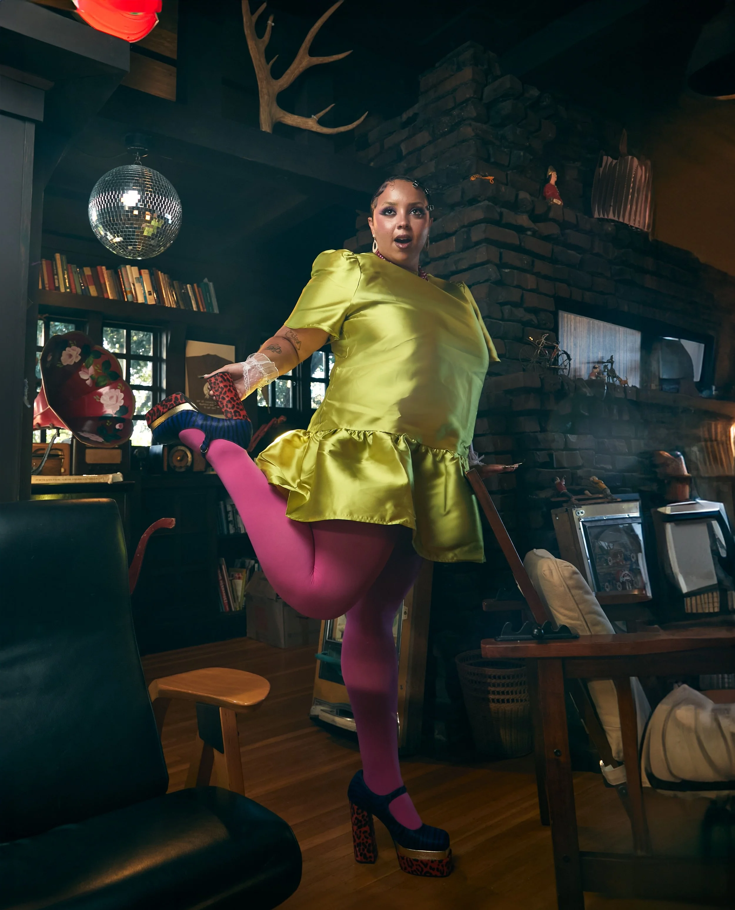 A woman in a bright yellow satin dress and pink tights wearing leopard print platform high heels, posing indoors with her mouth open, with a dark brick fireplace, bookshelves, and a reflective disco ball hanging from the ceiling in the background.