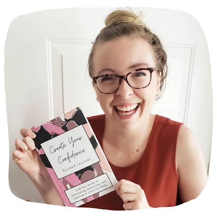 A young woman smiling and holding a book titled 'Create Your Confidence'. She is wearing glasses and a sleeveless top, standing in front of a white door.