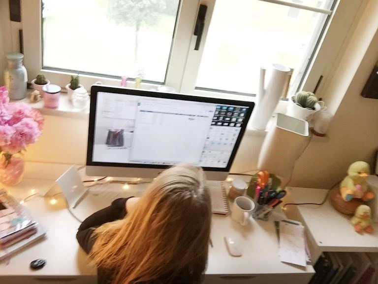 A person with long blonde hair working at a cluttered desk by a window, with a computer monitor, coffee mug, pens, papers, and decorative items.