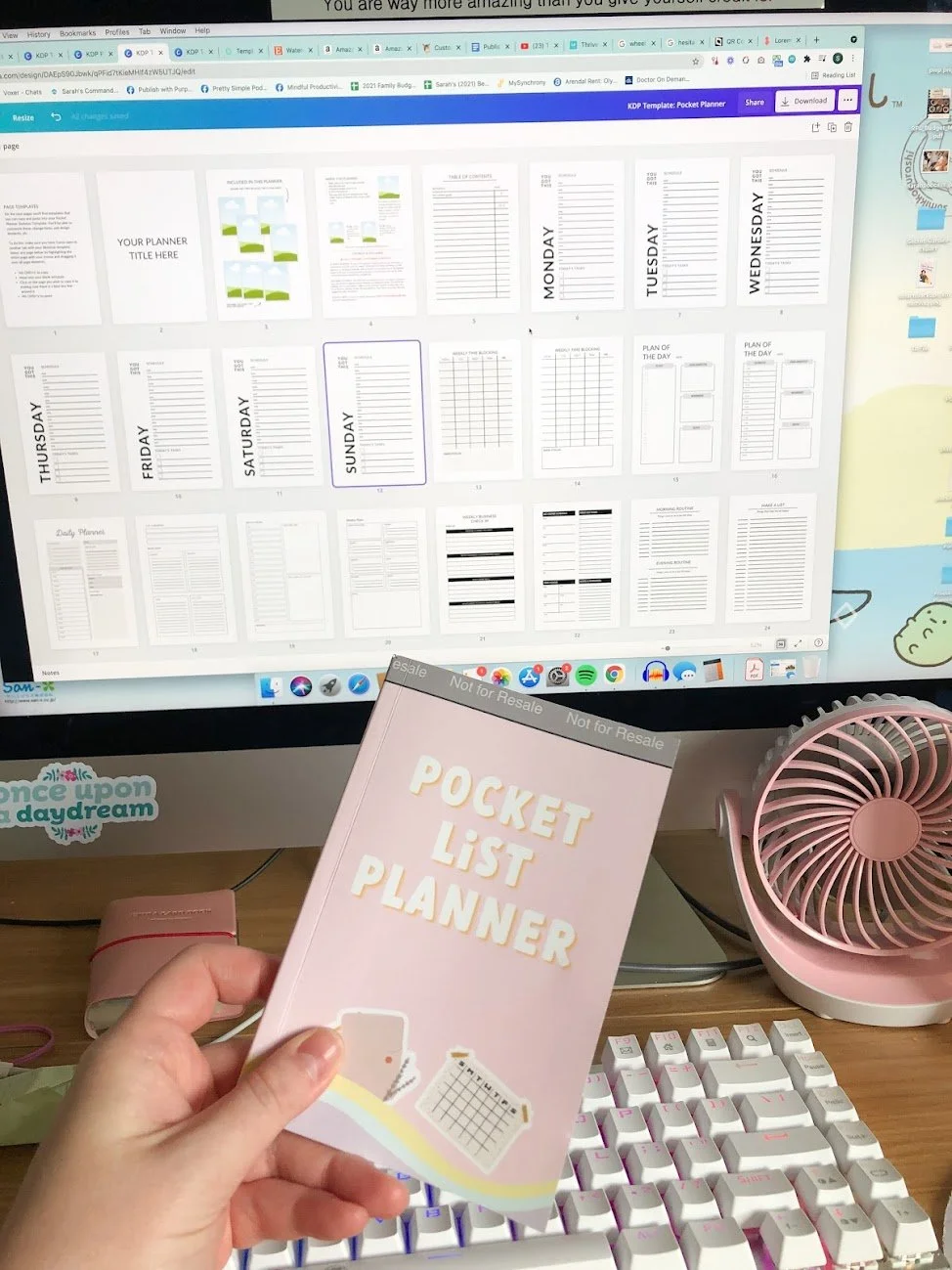 A person holding a pink pocket list planner in front of a computer monitor displaying a digital planner template with weekly and daily layouts. The workspace includes a pink fan, a white keyboard, and various small objects on the desk.