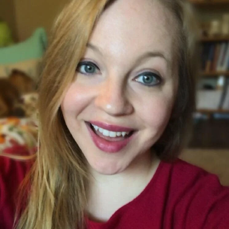 Close-up of a woman with long red hair, blue eyes, and a wide smile, wearing a red top, in a cozy indoor setting with a blurry background and a cat lying on a blanket.