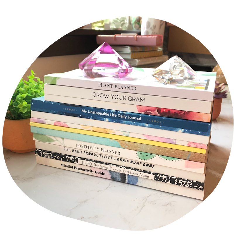 Stack of books including plant planner, grow your gram, my unstoppable life daily journal, positivity planner, the daily productivity & brain dump book, the 90-day novel planner, and mindful productivity guide, with two pink and clear decorative gemstones on top, a small potted plant to the left, and various items in the background.