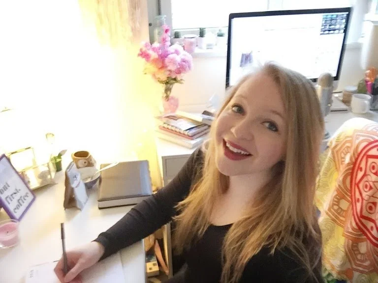 A woman sitting at a desk smiling at the camera, with a computer monitor, flowers, and books on the desk.