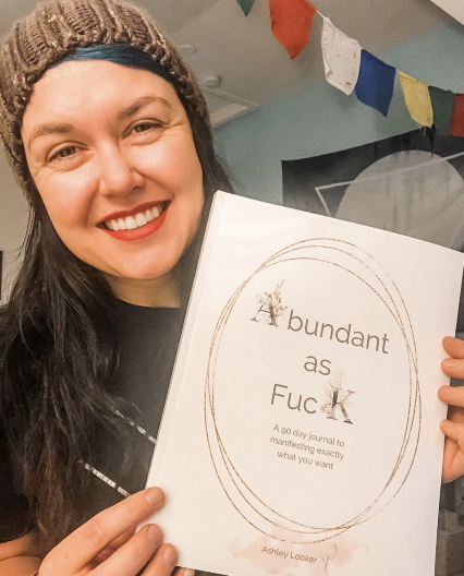 Smiling woman holding a book titled "abundant as F*** K" with colorful flags hanging in the background.