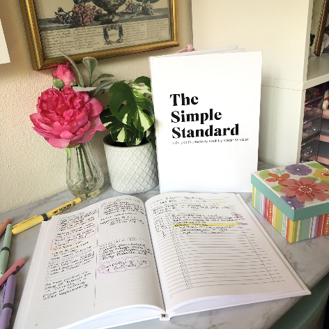 Open notebook with handwritten notes, pink and yellow highlighters, a pink flower in a glass vase, a white potted plant, a book titled 'The Simple Standard,' and a colorful box on a desk.