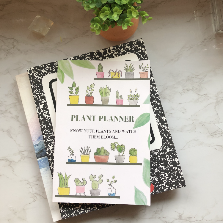 Plant planner book on top of a black and white patterned notebook, with a potted plant in a terracotta pot and a small candle on a light-colored marble surface.