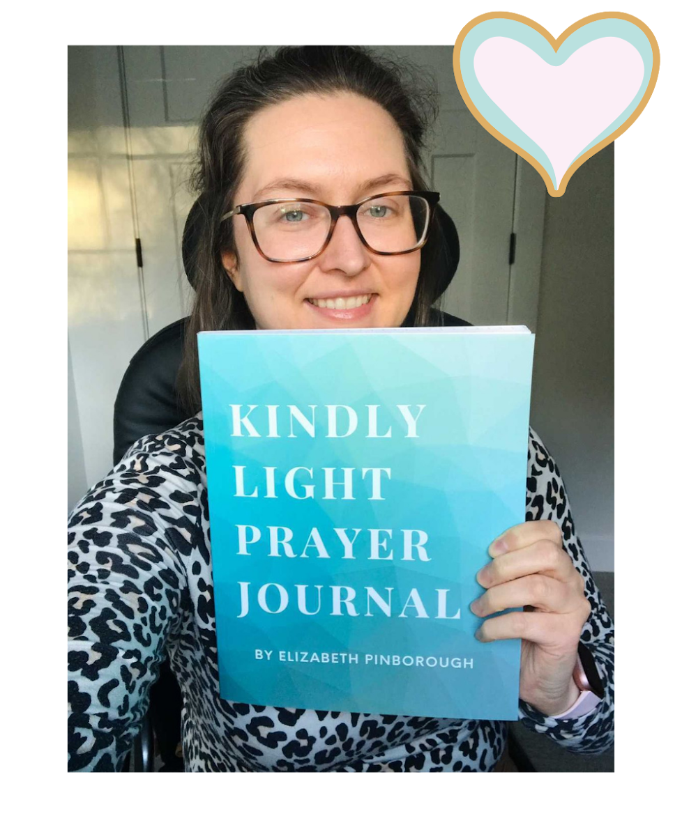 A woman with glasses and long brown hair smiling and holding a blue book titled 'Kindly Light Prayer Journal' by Elizabeth Pinborough. She is sitting in a black chair in front of a background with cabinets. There is a heart graphic in the top right corner of the image.