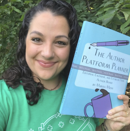 Woman with dark curly hair smiling outdoors, holding a blue signed planner titled 'The Author Platform Planner' with a purple latte mug illustration on the cover.