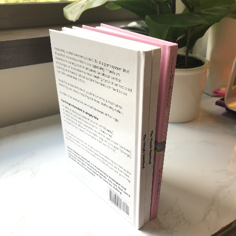 A stack of three books, with the top book open and facing away, on a white marble surface near a window and a potted plant.