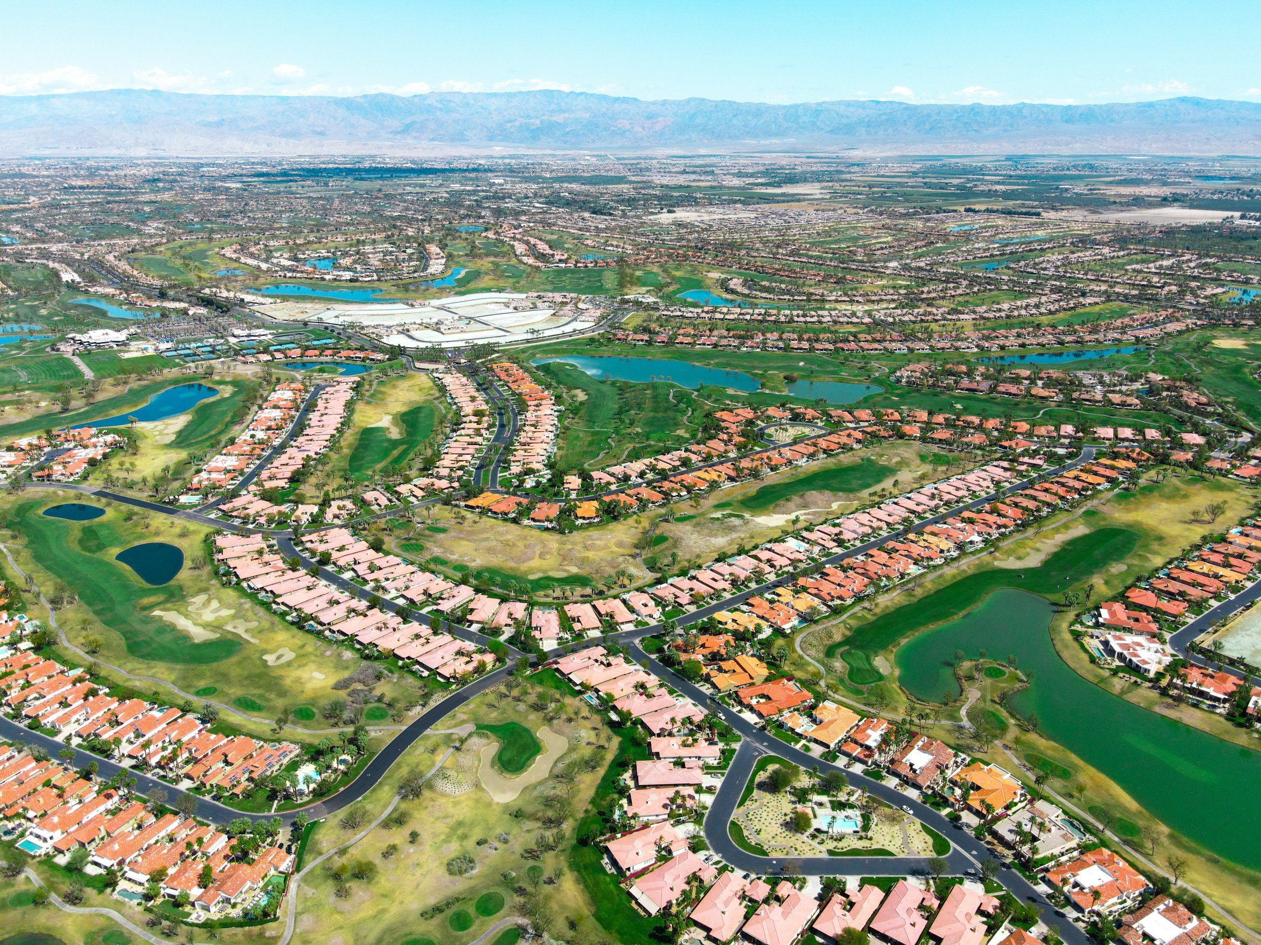 Aerial view of a suburban residential area with numerous houses, green lawns, small lakes, and a golf course, with mountains in the background.