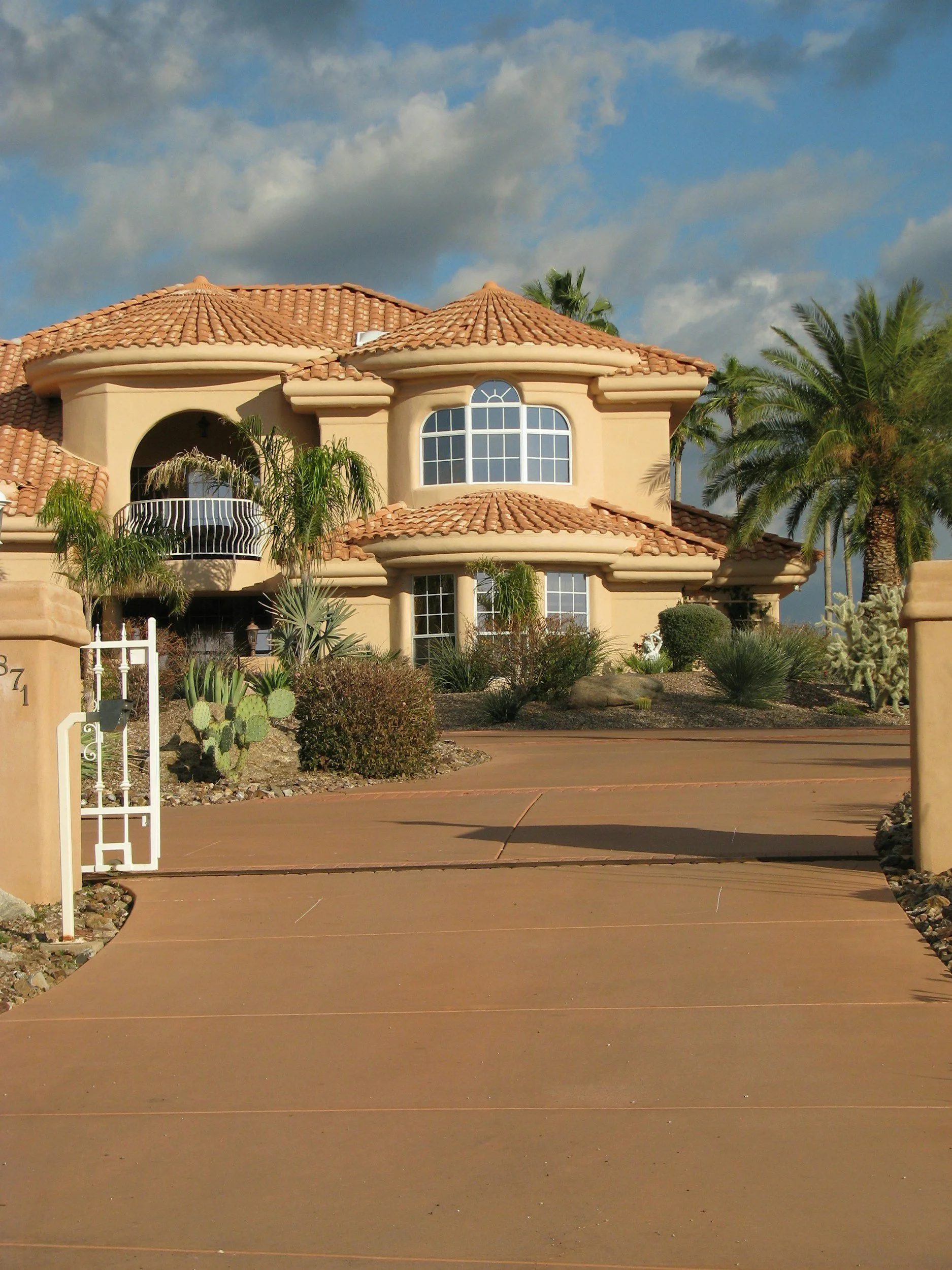 A large beige house with a red tile roof, tall arched window, and a balcony, surrounded by desert plants and palm trees. The driveway is curved and paved.