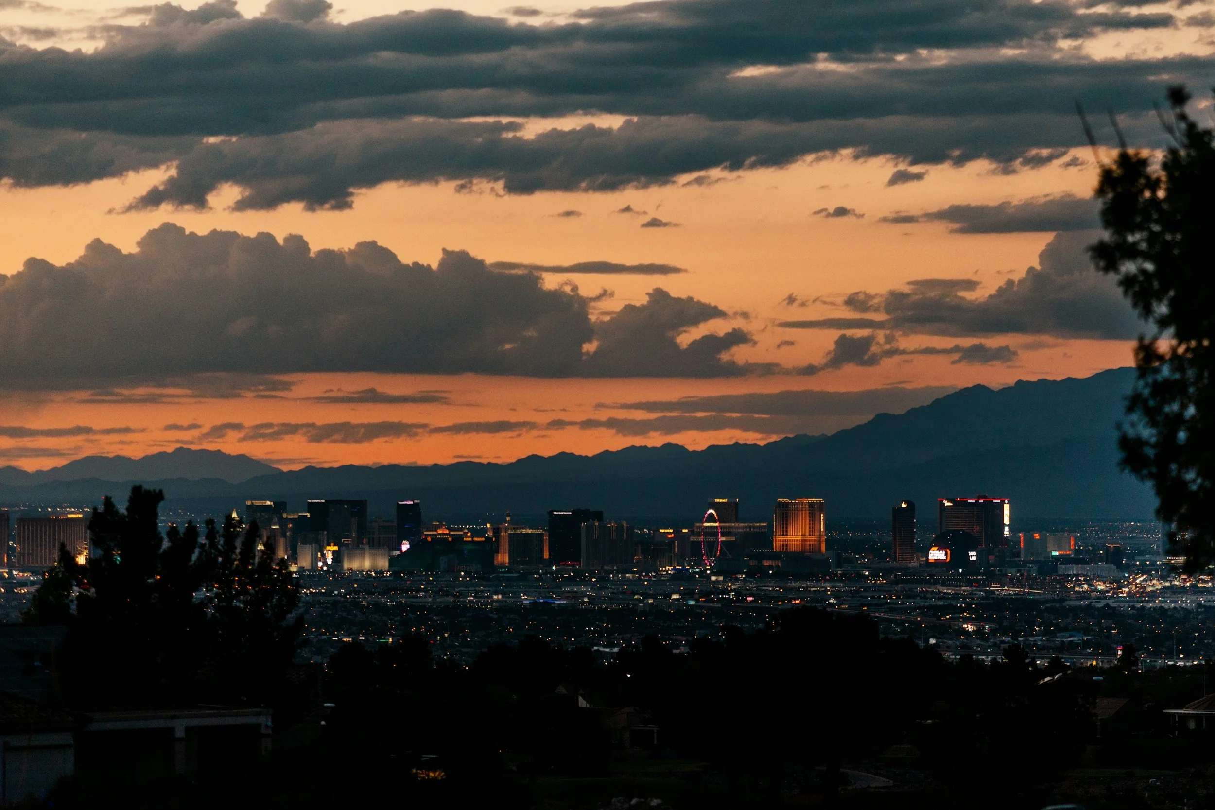 City skyline at dusk with mountains in the background, illuminated buildings, and dark clouds in the sky.