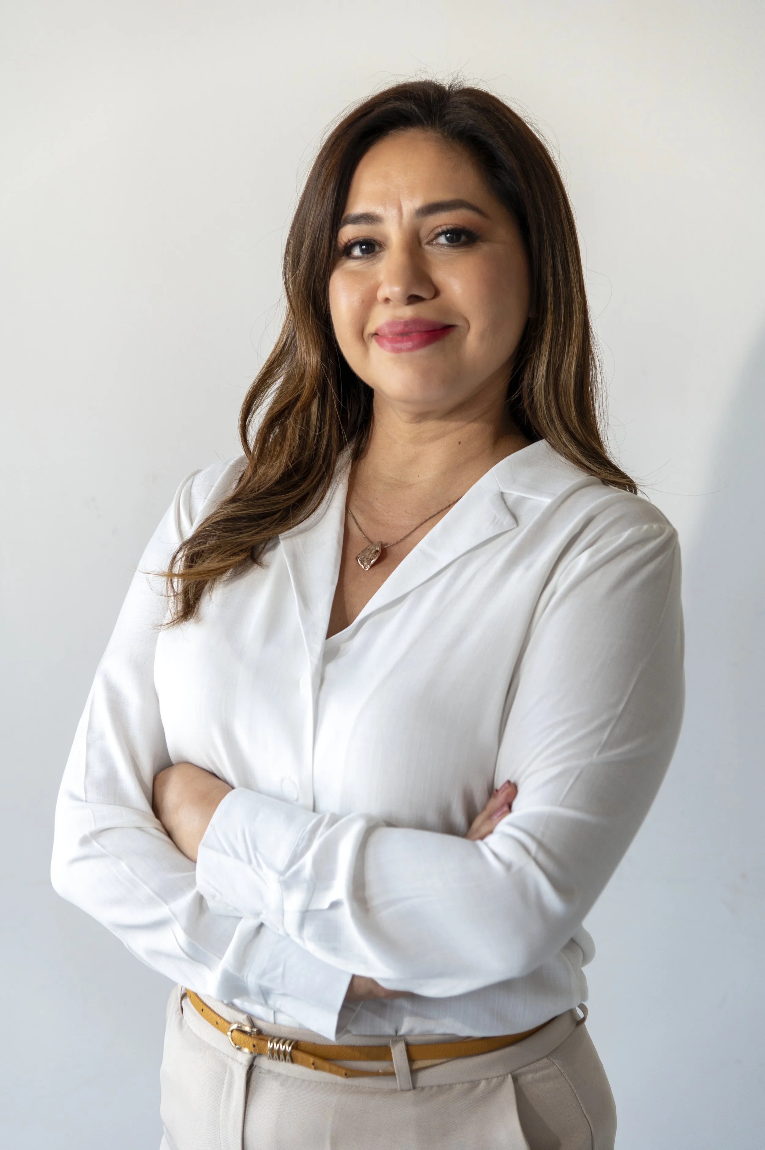 A woman with long brown hair and light skin, wearing a white blouse with rolled-up sleeves, standing against a plain light background with her arms crossed, smiling softly.