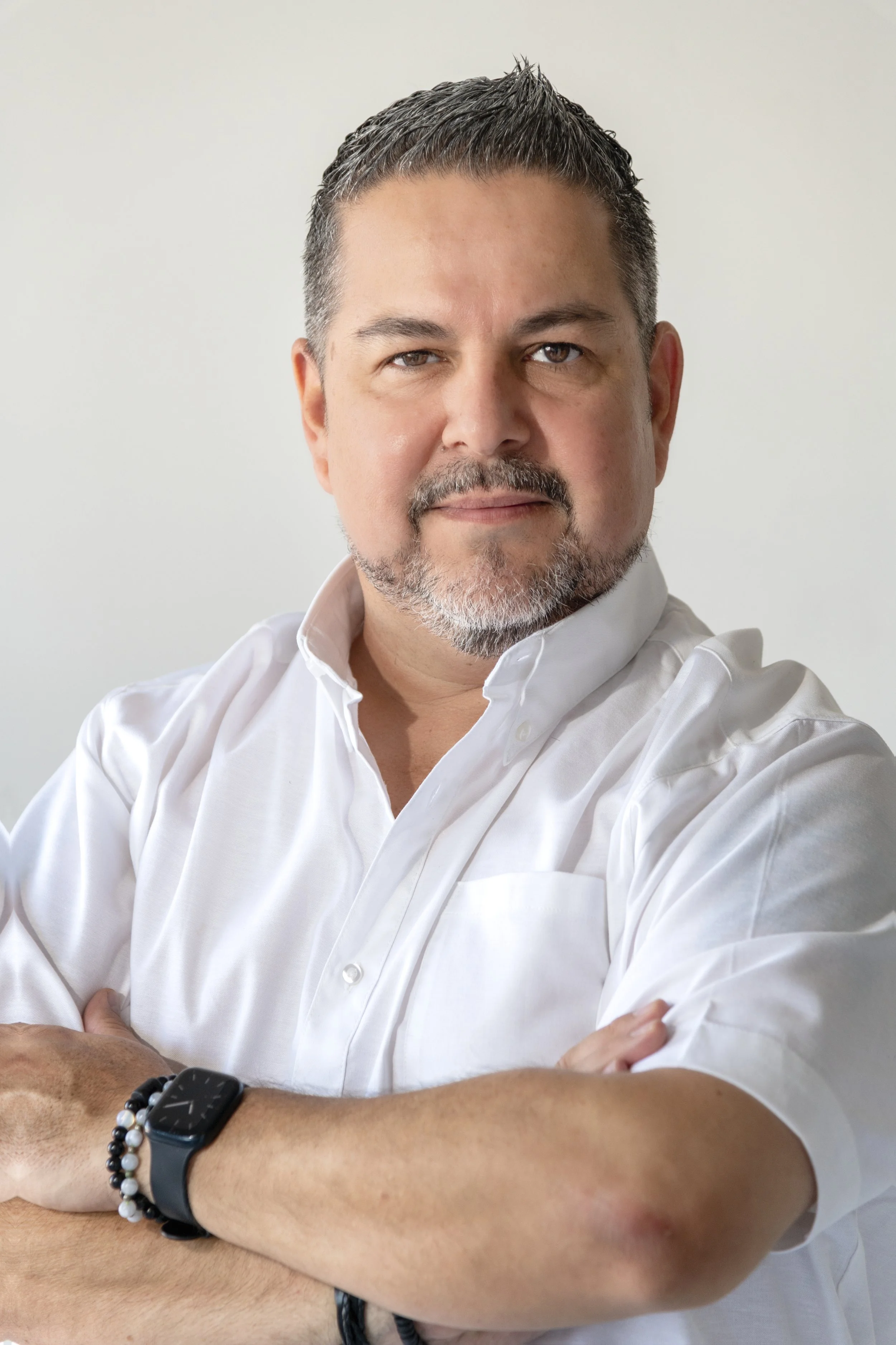 A man with dark hair, gray beard, and mustache, wearing a white dress shirt, with arms crossed, and a smartwatch and beaded bracelet on his left wrist, looking at the camera against a plain light background.