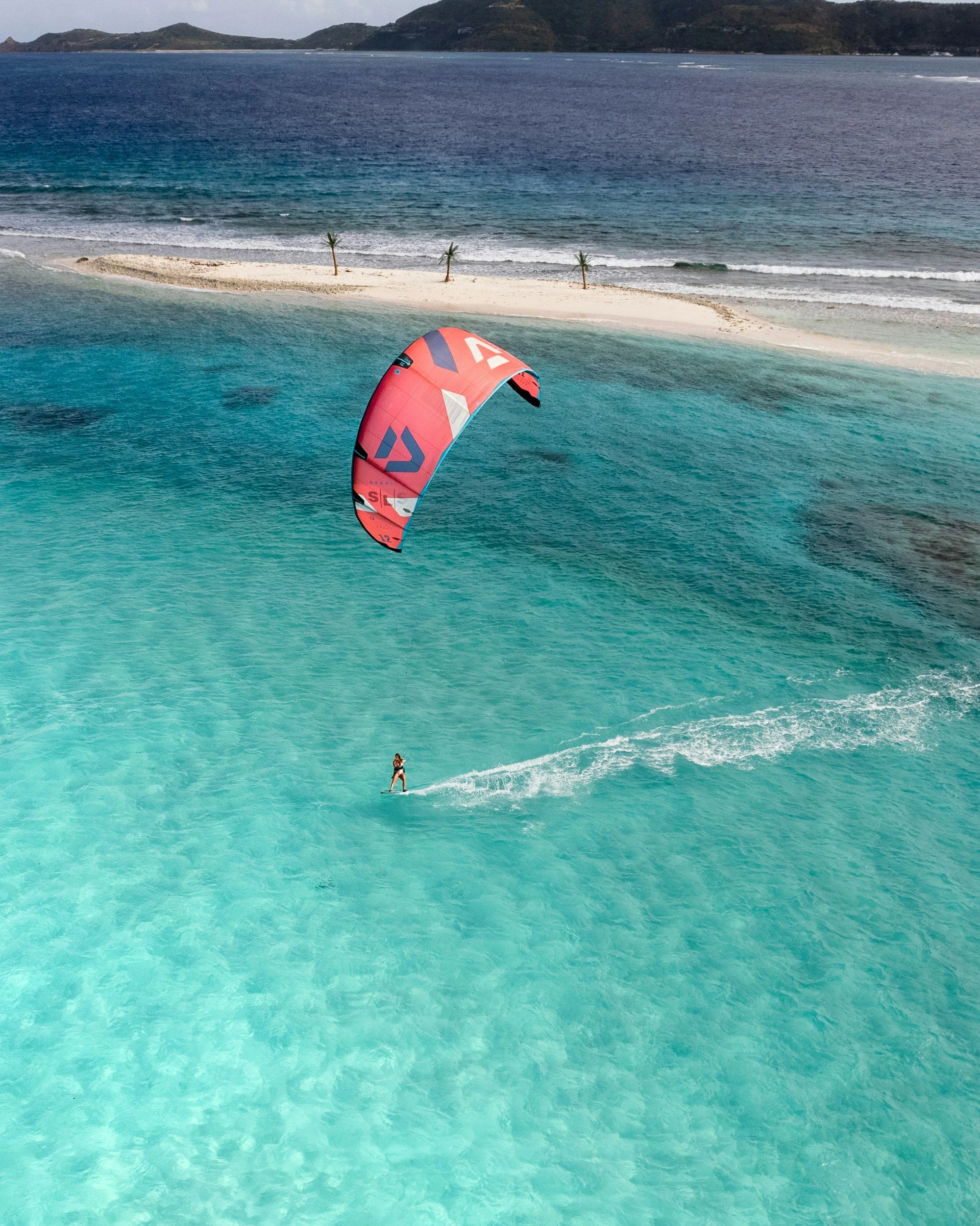 A person kite surfing on clear blue water near a sandy beach with small palm trees and an island in the background.
