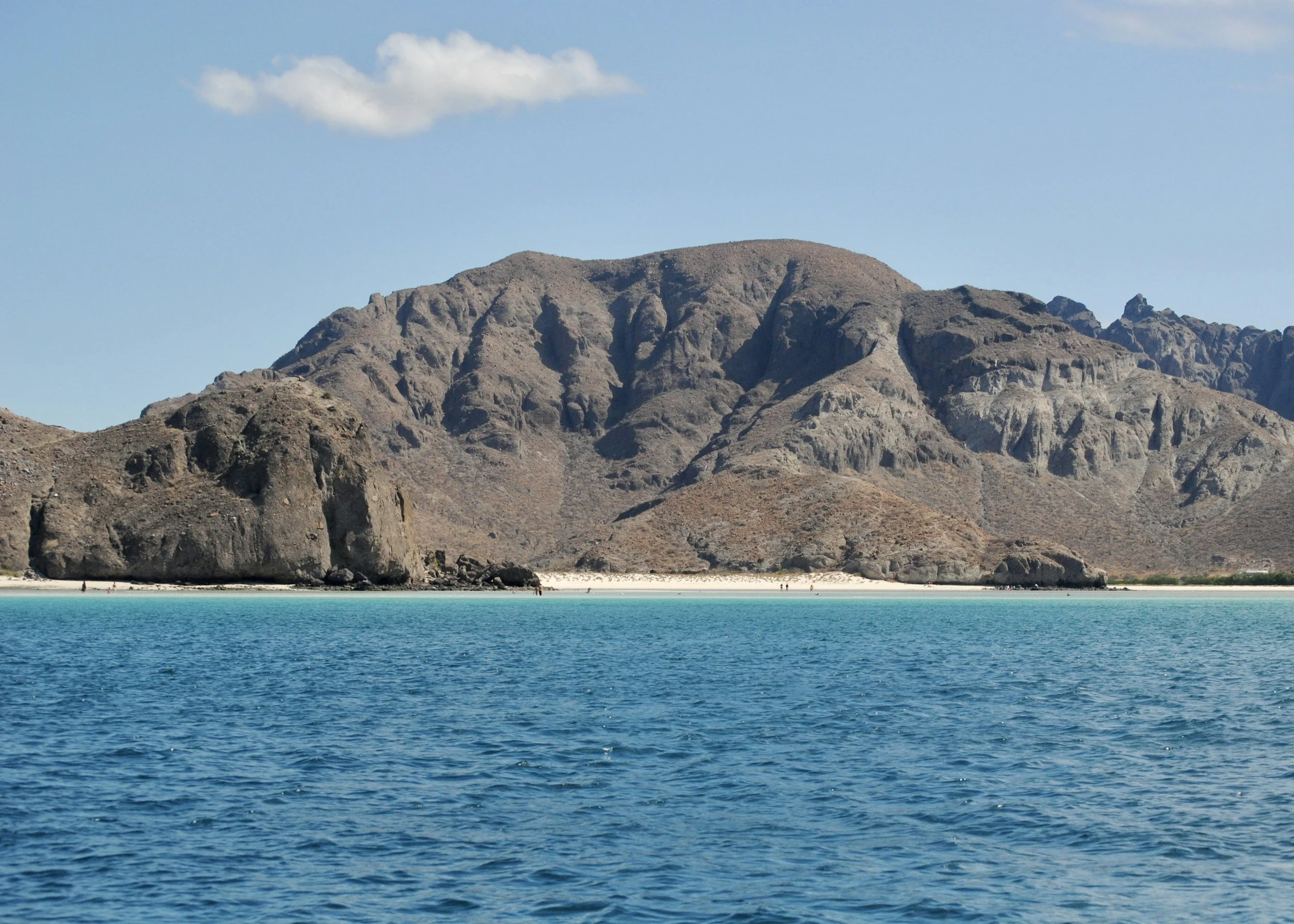 A large rocky mountain by the water with a sandy beach and a few people walking along the shoreline under a partly cloudy sky.