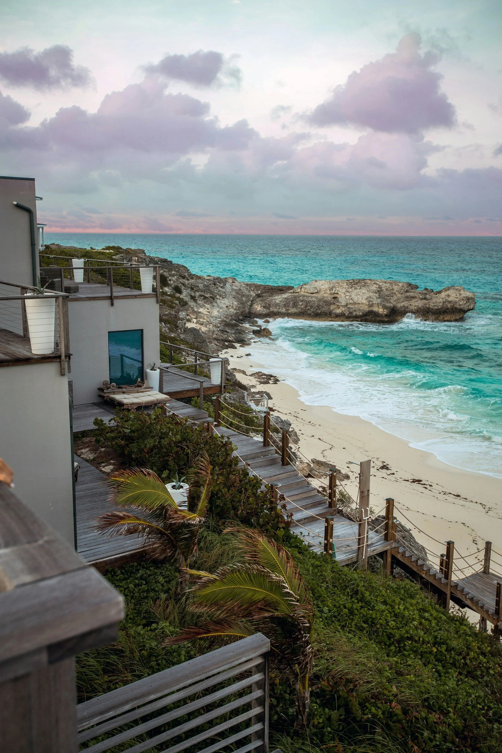 View of a beach with rocky shoreline, turquoise ocean waves, and a cloudy sky, seen from a multi-level deck with plants and railing.