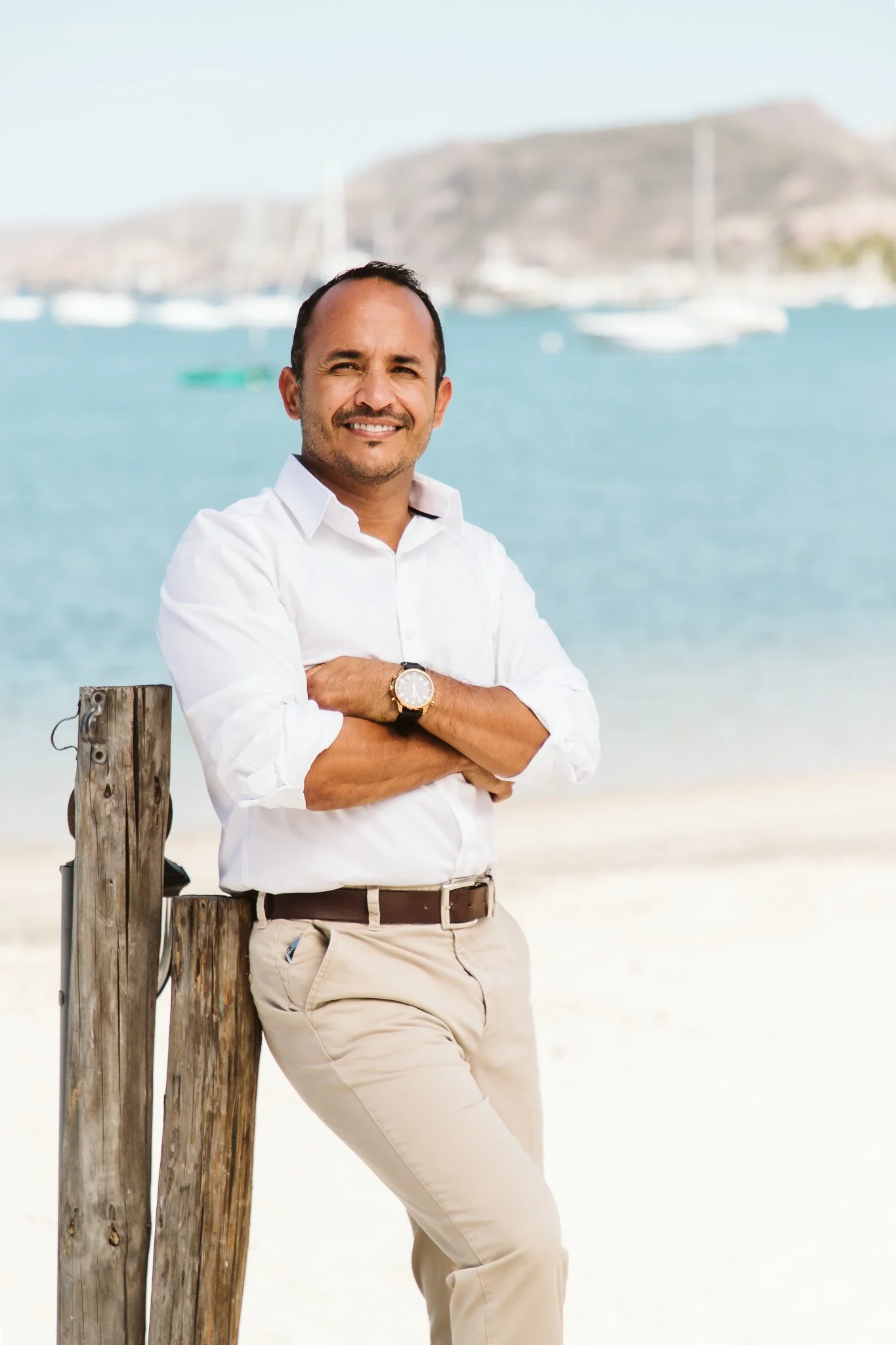 A man in a white shirt and beige pants standing outdoors near a wooden post with a marina and sailboats in the background.