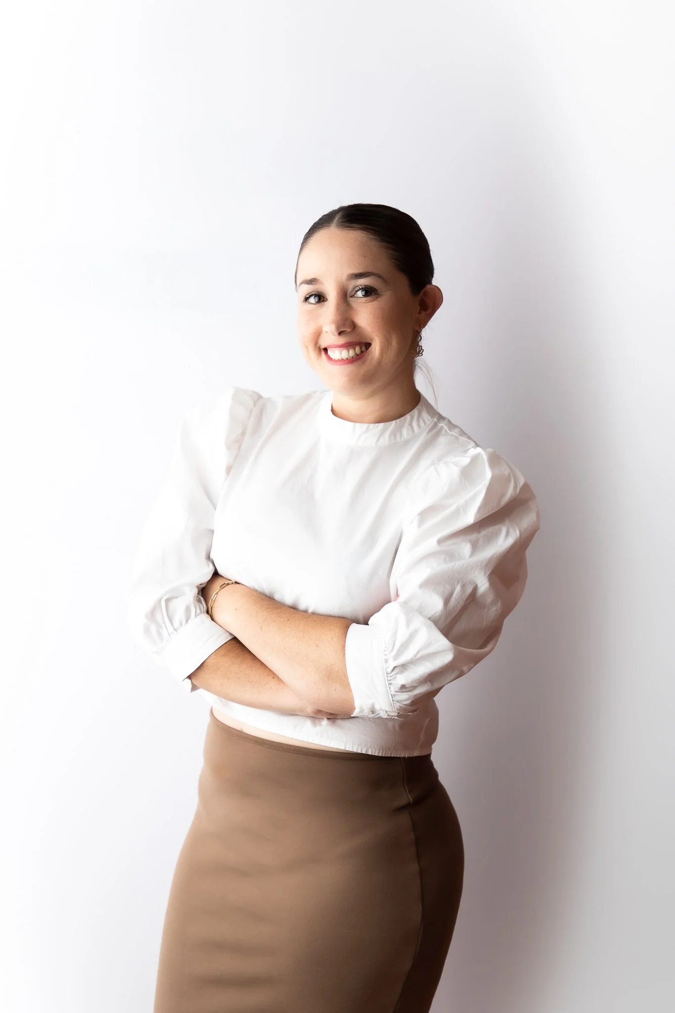 A young woman with dark hair tied back, smiling and crossing her arms. She is wearing a white blouse with puffed sleeves and a tan skirt, standing against a plain white background.