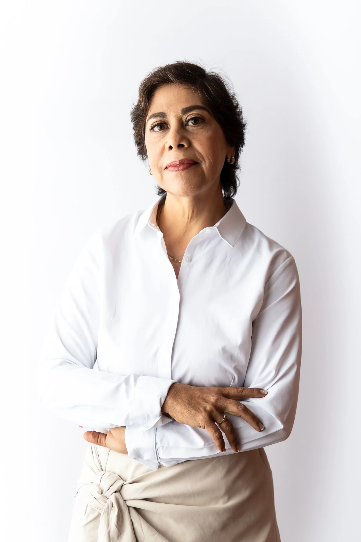 A middle-aged woman with short, dark hair, wearing a white button-up shirt, poses with her arms crossed against a plain white background.