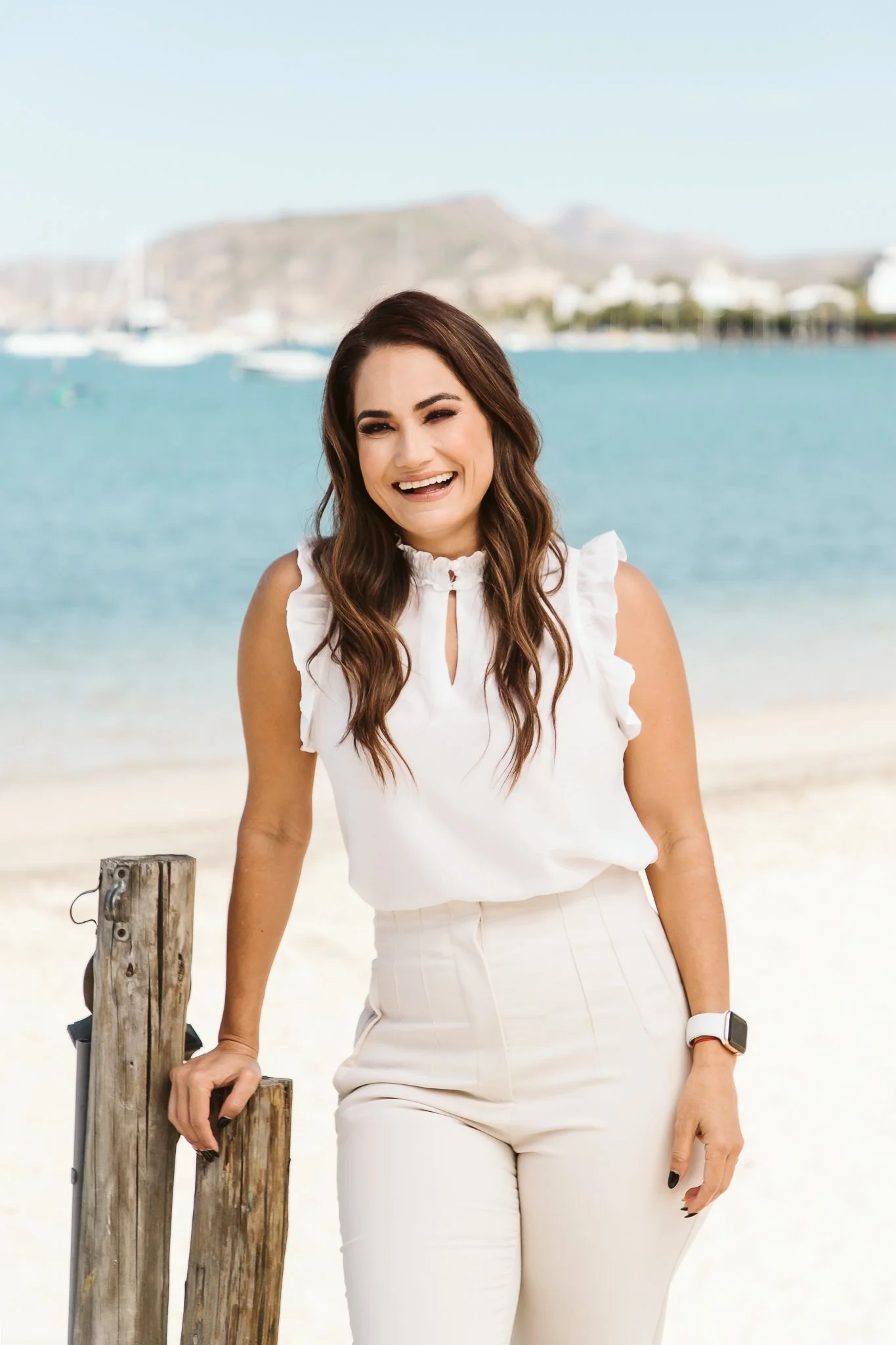 Woman with long brown hair smiling at the camera, wearing a white sleeveless top and white pants, standing next to a weathered wooden post at the beach with water and a mountain in the background.