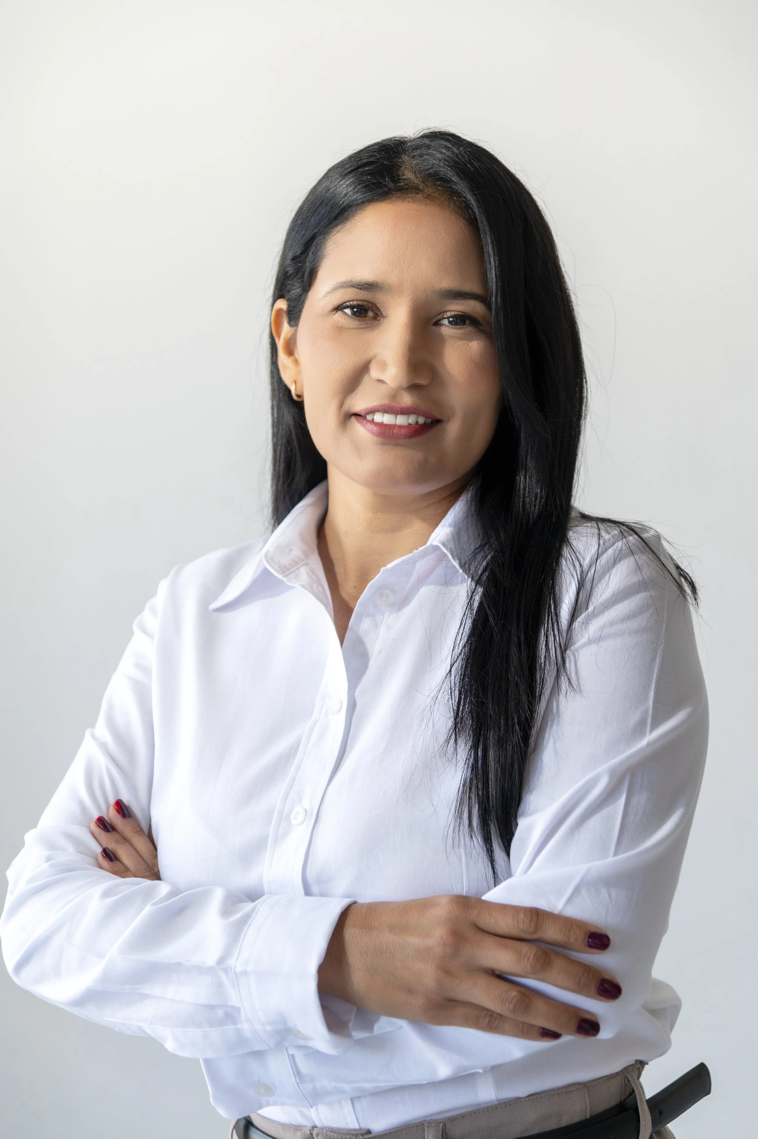 A woman with long black hair, wearing a white button-up shirt, standing with arms crossed, smiling, against a plain white background.