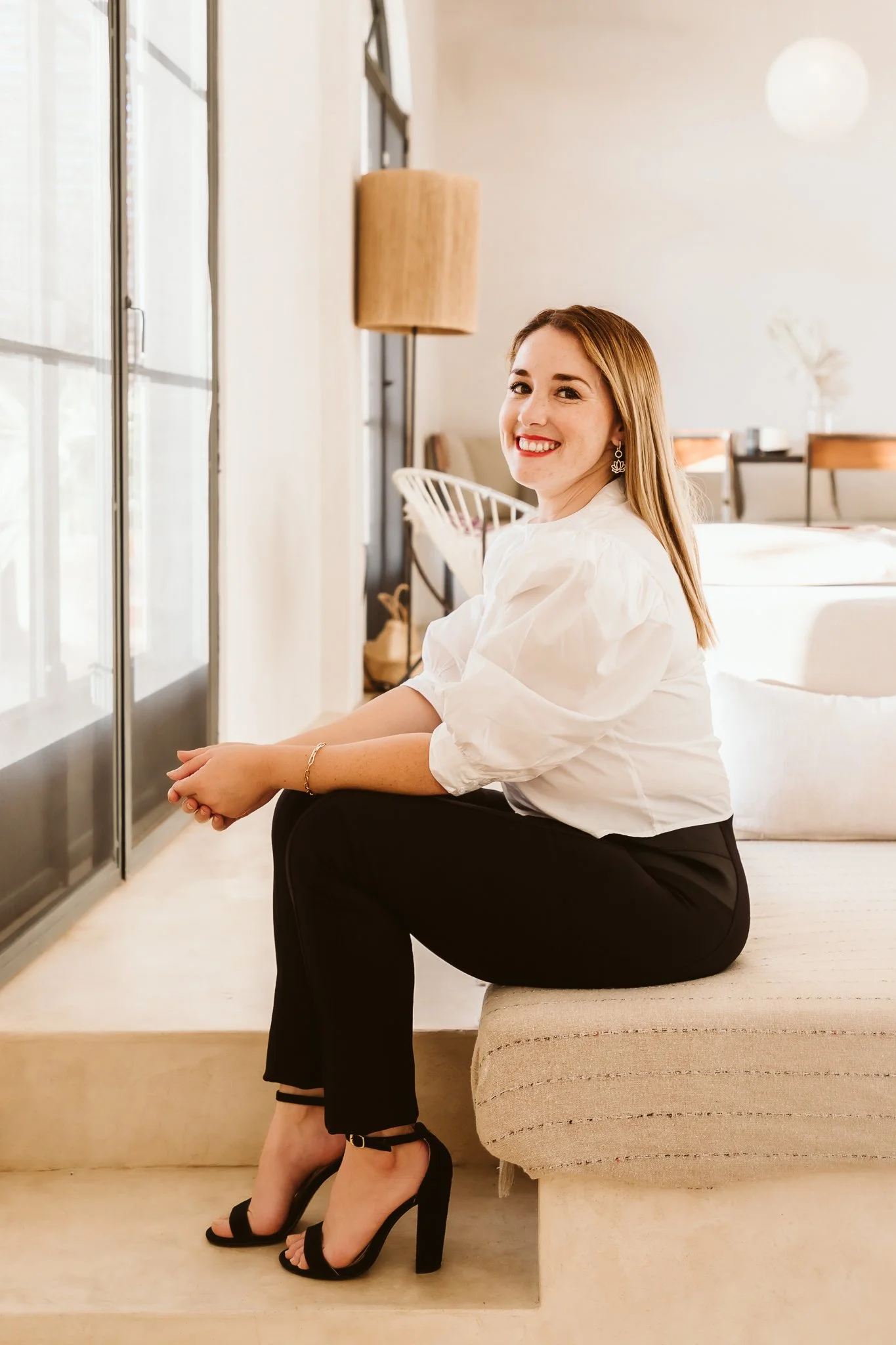 A smiling woman with long blonde hair, wearing a white blouse, black pants, and black high-heeled sandals, sitting on a beige bench in a well-lit modern room with large windows and minimalist decor.