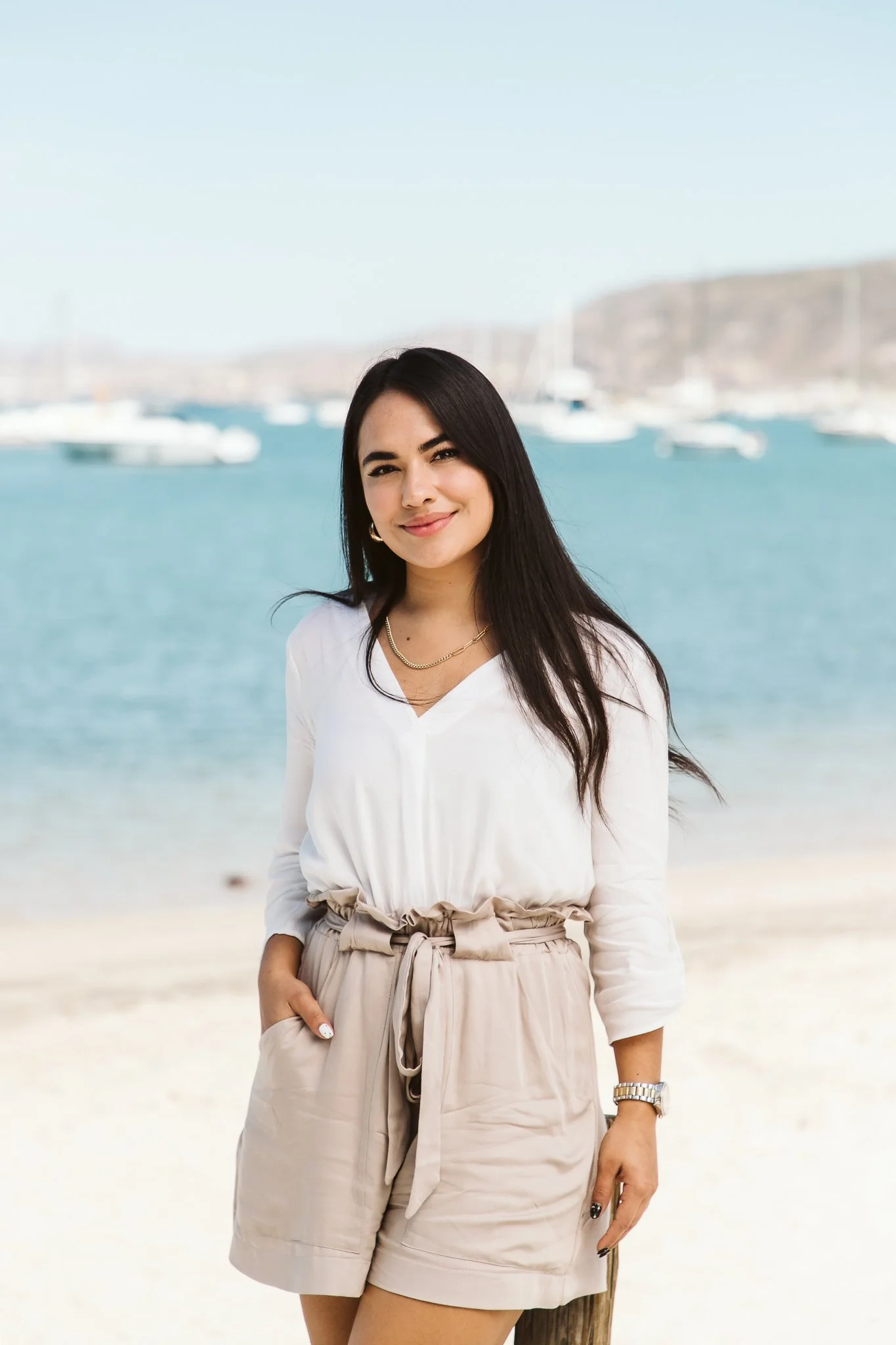 A woman with long dark hair smiling at the camera, dressed in a white blouse and beige high-waisted shorts, standing on a beach with sailboats and a hill in the background.