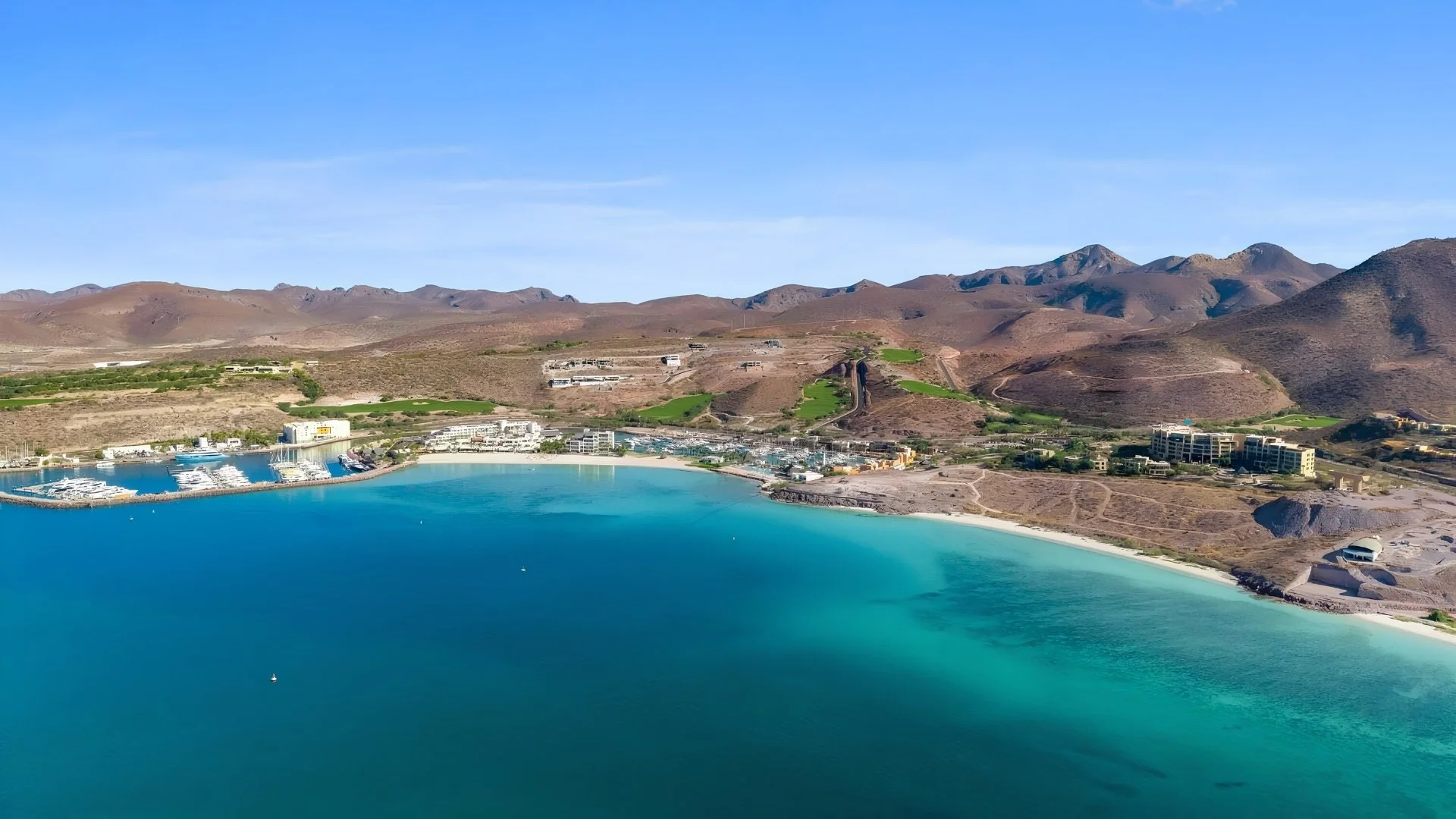 Aerial view of a coastal area with a beach, marina, and buildings against mountains under a clear blue sky.
