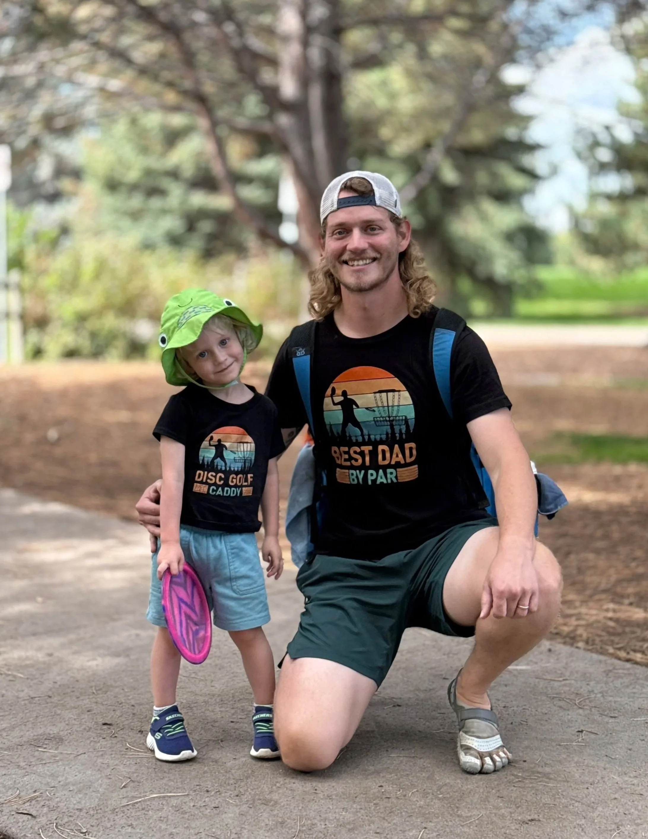 A man and a young girl posing outdoors in a park, with trees in the background. The girl is wearing a green hat with eyes and a mouth, a black t-shirt with a graphic and text, and light blue shorts, holding a pink and purple frisbee. The man is wearing a gray backwards cap, a black t-shirt with a colorful graphic and text, green shorts, hiking sandals, and has a backpack. The man has one knee on the ground and is smiling at the camera.